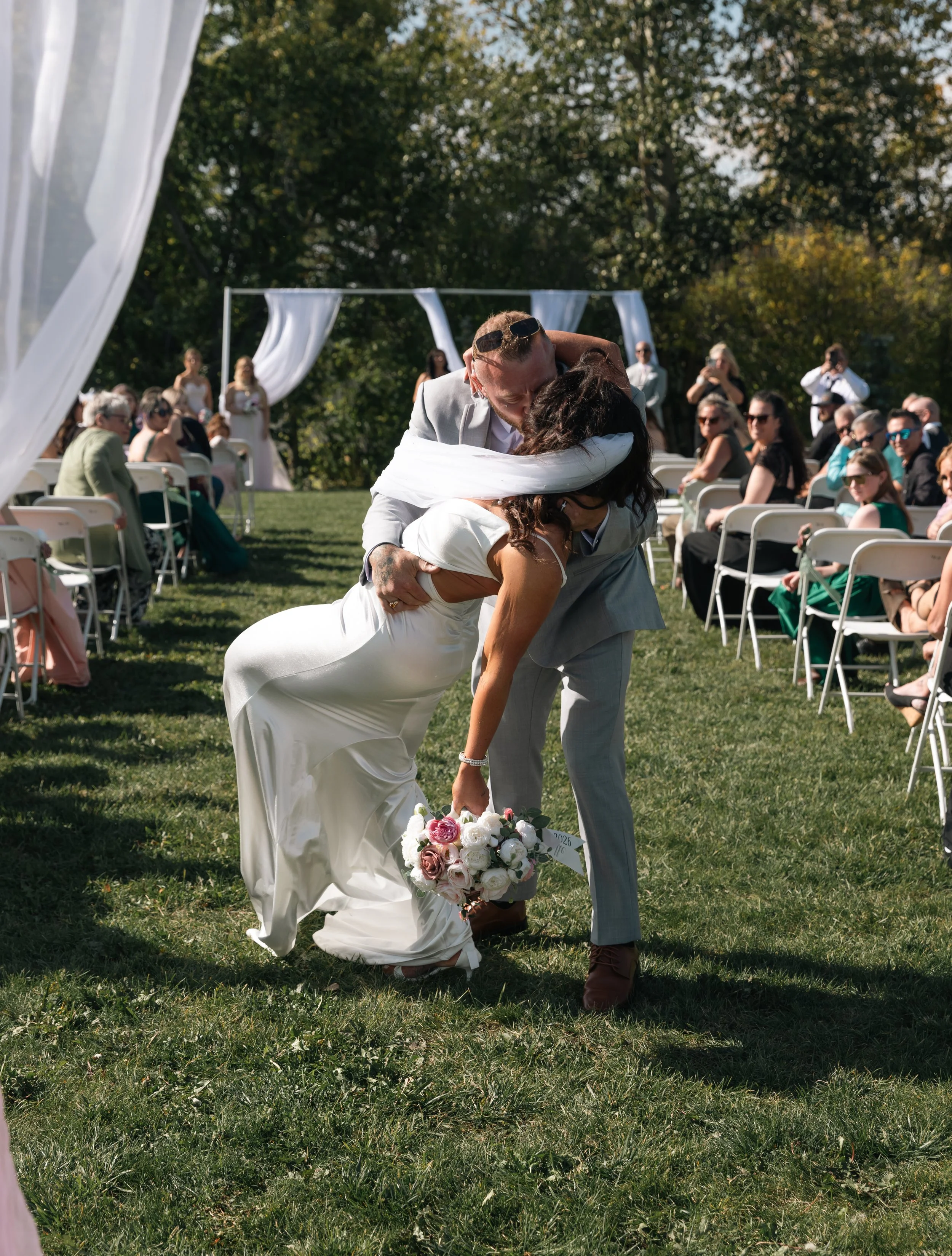A wedding scene outdoors with a bride and groom sharing a kiss, the bride is bending down to pick up her bouquet, while the groom leans over her. Guests are seated in chairs in the background, some watching and others talking. Decorative white fabric