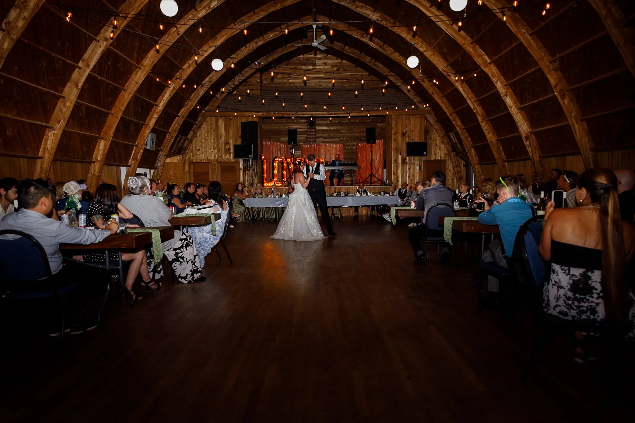 A bride and groom are dancing in a rustic wooden barn decorated for a wedding reception, with guests seated at tables around them and string lights hanging from the ceiling.