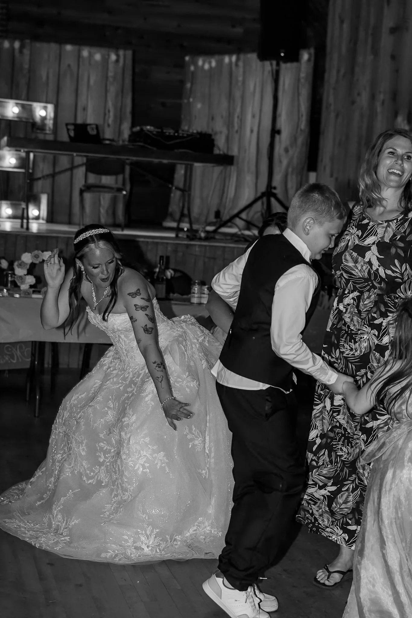 A wedding reception scene with a bride in a lace wedding gown dancing with a young boy in a vest and dress shirt. An older woman in a floral dress is dancing nearby. The background features a wooden wall, a DJ setup, and a table with flowers and bottles.