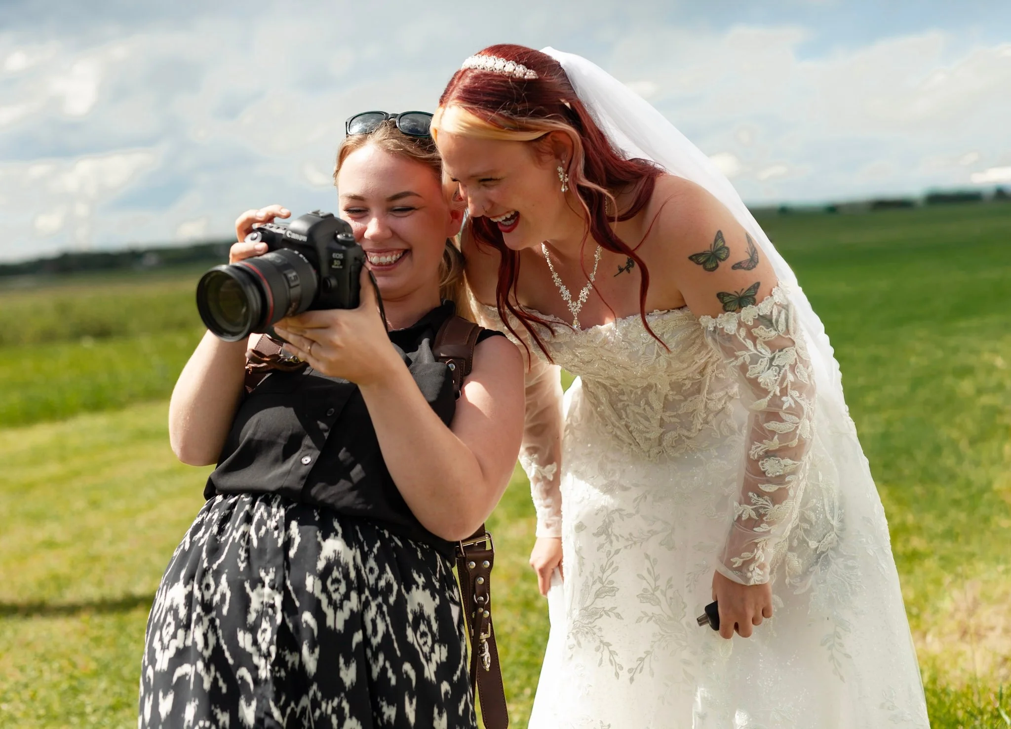 Two women at a wedding, one in a wedding dress with tattoos and the other in a black and white patterned dress, looking at a camera and smiling outdoors on a cloudy day.