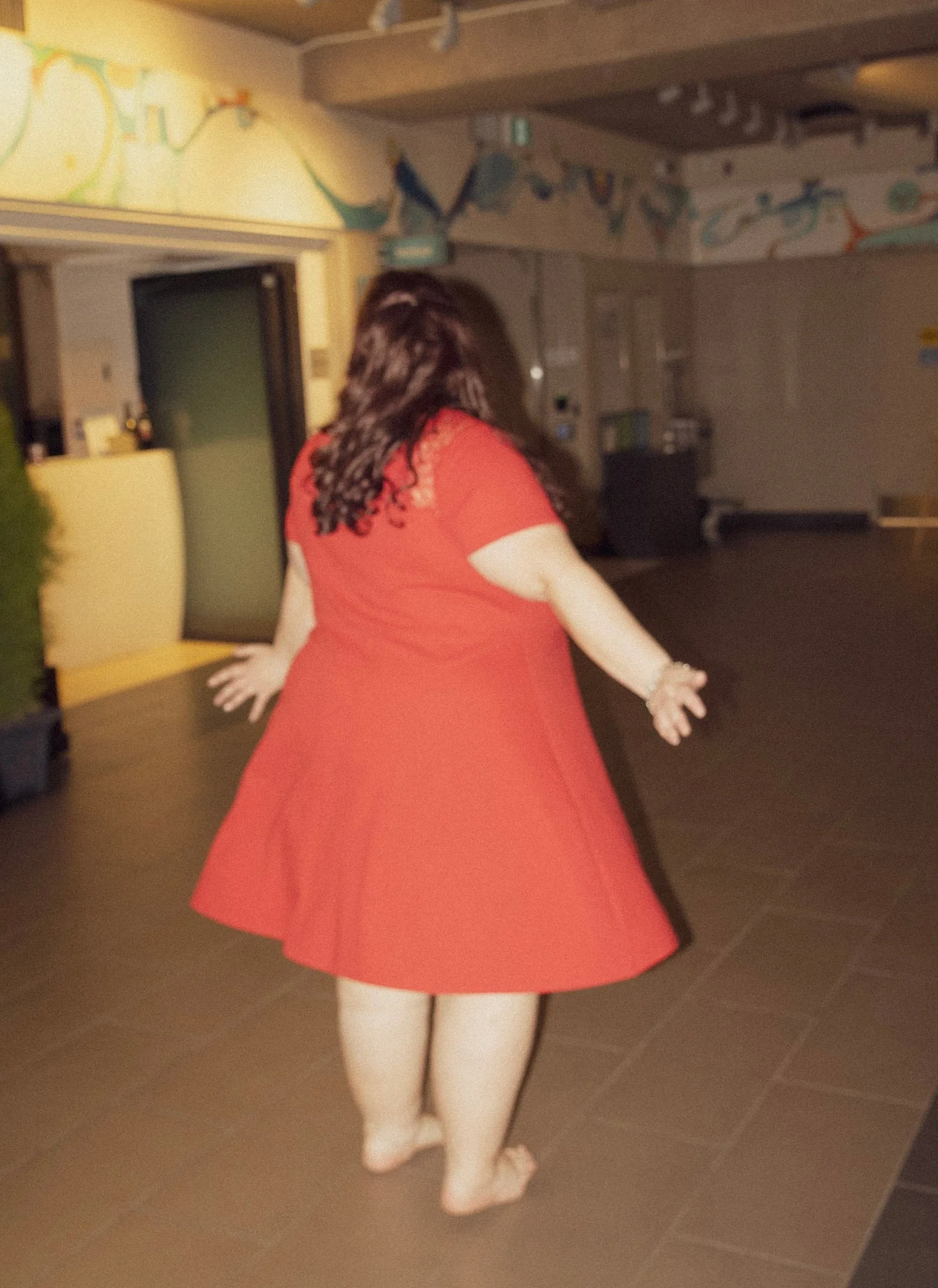 A person wearing a red dress with curly hair, standing with arms outstretched in an indoor space with beige toned walls and a colorful mural arching near the ceiling.