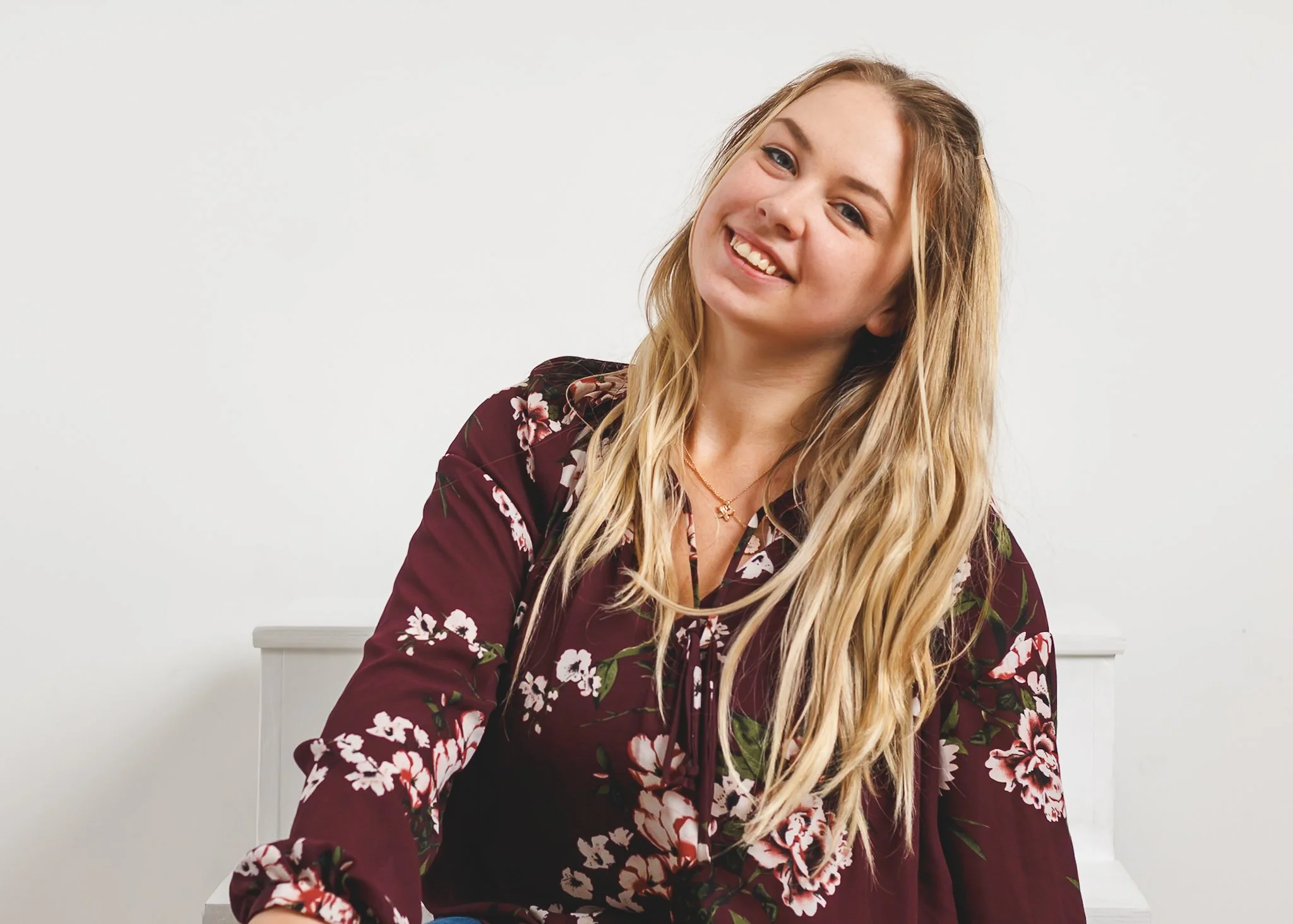 Portrait of a young woman with long blonde hair, smiling, wearing a maroon floral blouse, sitting against a plain white background.