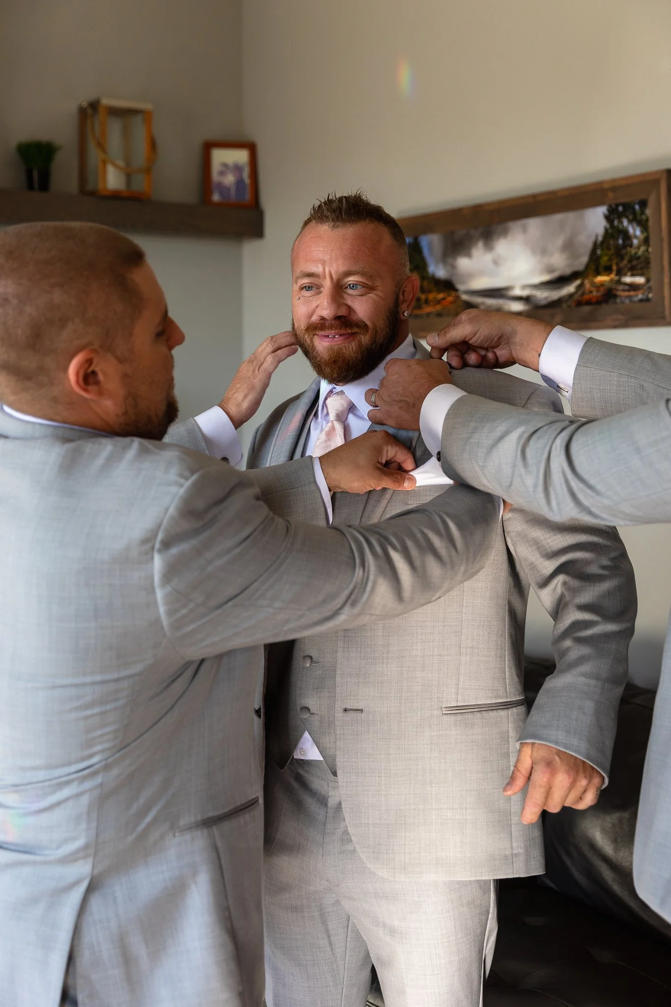 Three men in suits are helping a man in a suit with a tie, smiling, in what appears to be a living room or hallway with framed pictures and artwork on the wall.