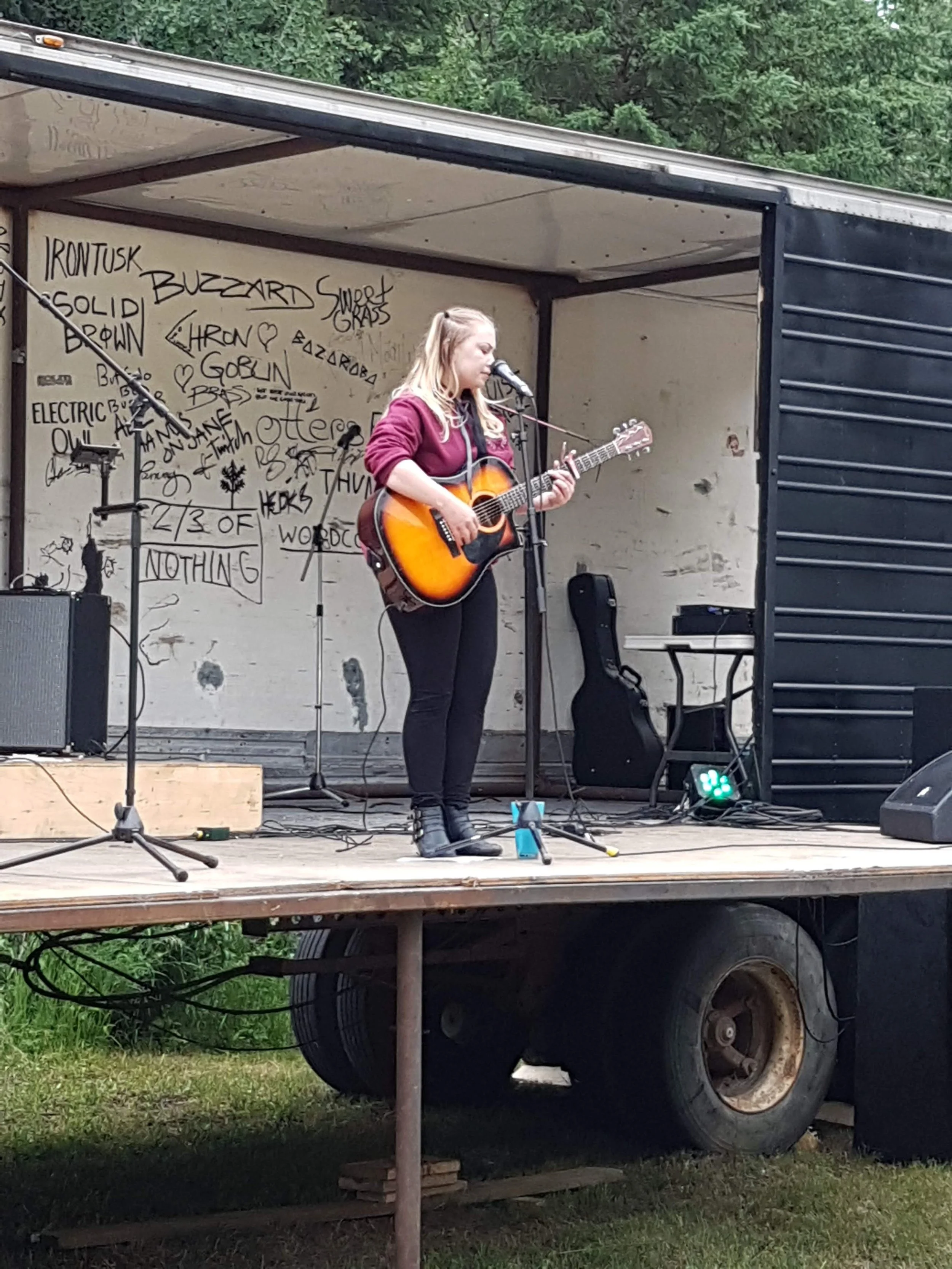A young woman with long blonde hair playing an acoustic guitar and singing into a microphone on an outdoor stage. The stage is set up in the back of a trailer truck with graffiti on the interior wall. There is equipment including speakers, a microphone, and a keyboard on the stage.