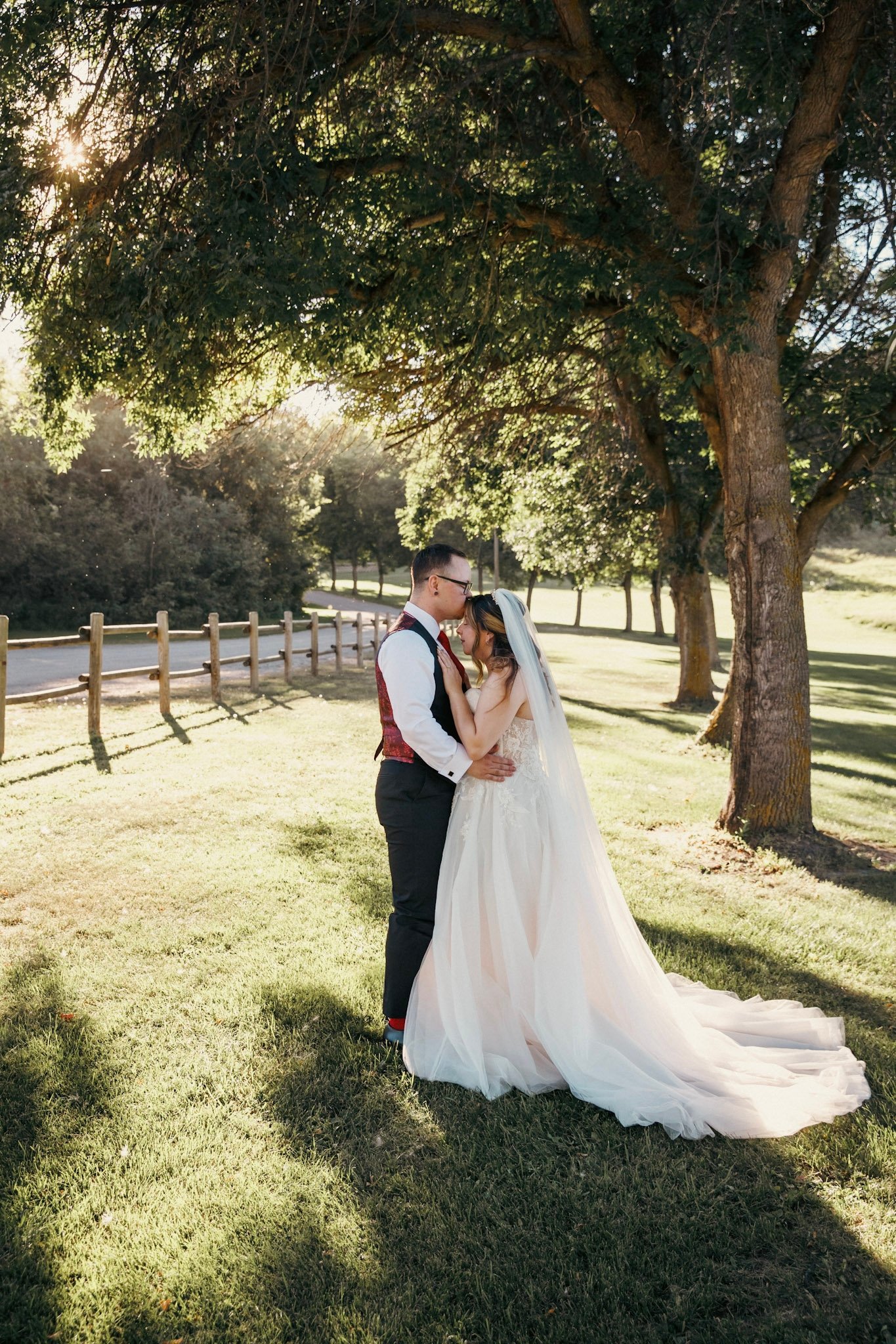 A bride and groom standing close together under a large tree in a park, with sunlight filtering through the leaves. The bride is wearing a white wedding gown with a long train and veil, while the groom is dressed in a black vest, white shirt, and bla