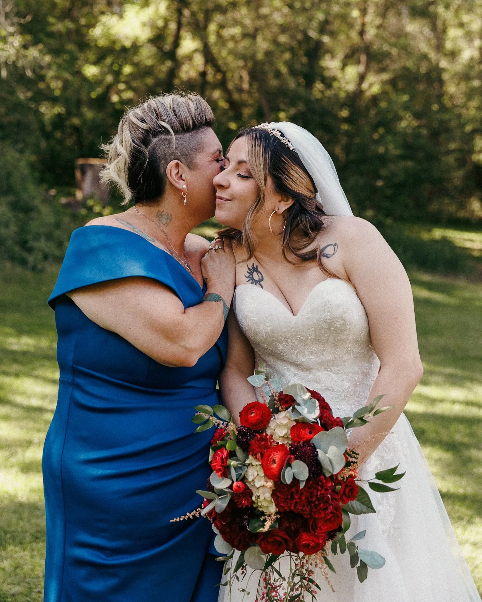 Two women, one dressed in a wedding gown and the other in a blue dress, sharing a kiss on the forehead outdoors with green trees and grass in the background. The bride holds a large bouquet of red, white, and green flowers.
