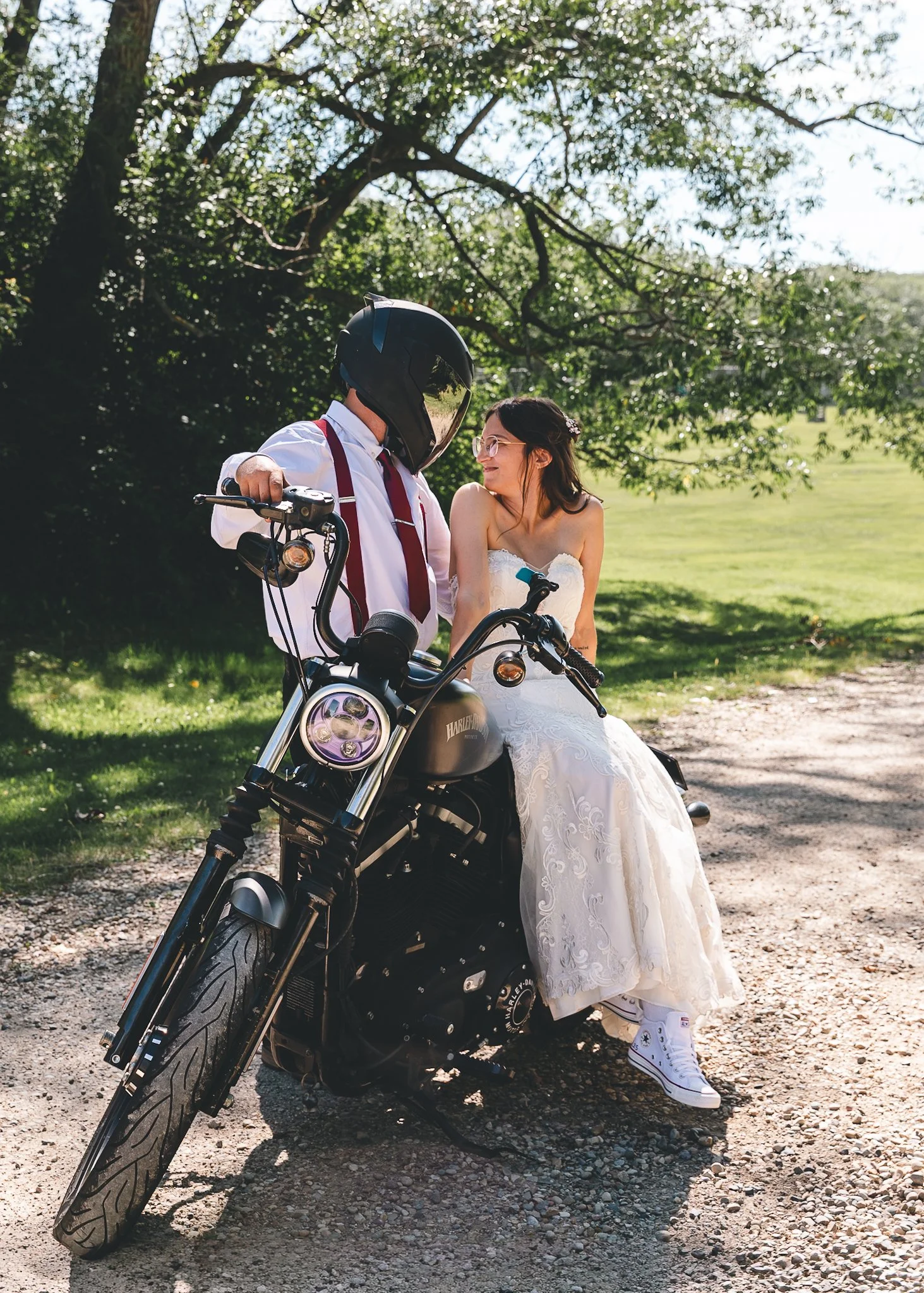 A woman in a white wedding dress and sneakers standing next to a man in a white shirt, red suspenders, and a motorcycle helmet, with a black Harley Davidson motorcycle outdoors in a park-like setting.