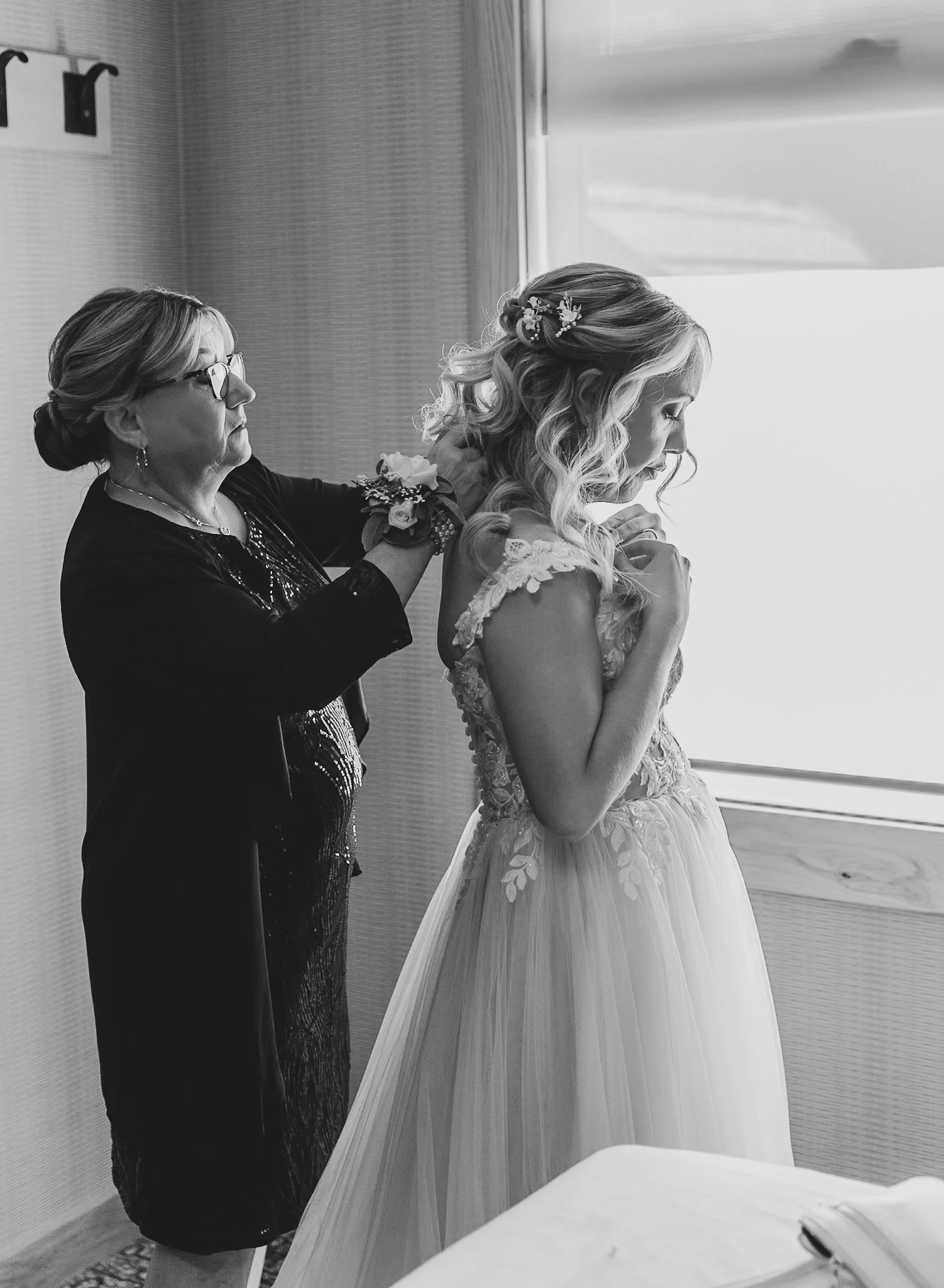 A bride with blonde curly hair and floral hair accessory, wearing a lace wedding gown, prays with hands clasped in front of a mirror while an older woman, possibly her mother or bridal attendant, adjusts her dress. The scene is set indoors near a win