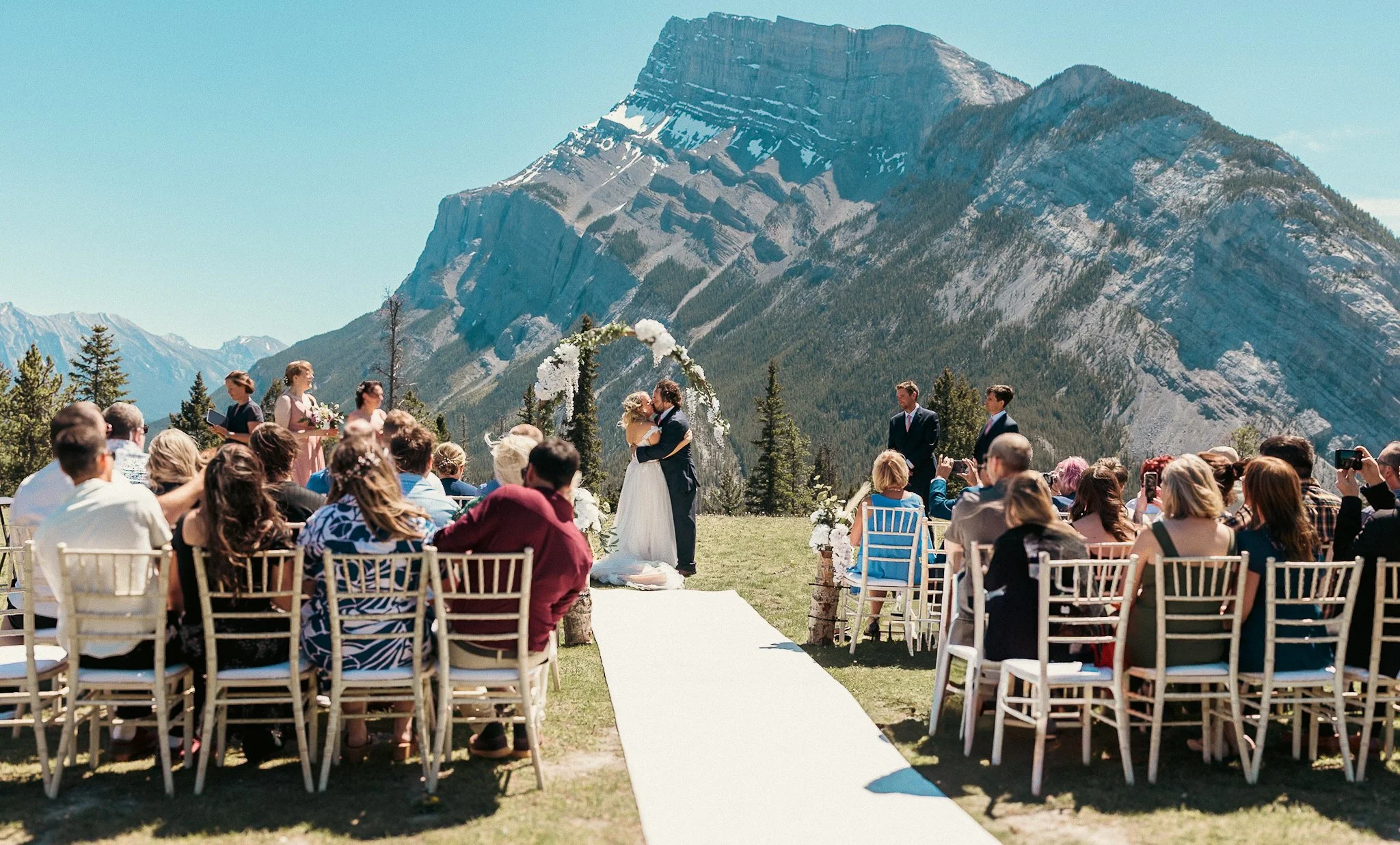 A wedding ceremony taking place at Tunnel Mountain in Banff. The bride and groom are standing under a floral arch, surrounded by guests seated on white chairs on a grassy field. The sky is clear and blue, indicating a sunny day.