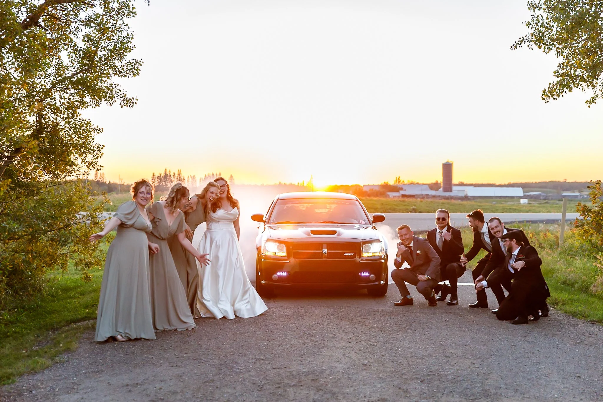 Group of people including women in dresses and men in suits posing around a black sports car on a rural road at sunset, with farm buildings and open fields in the background.
