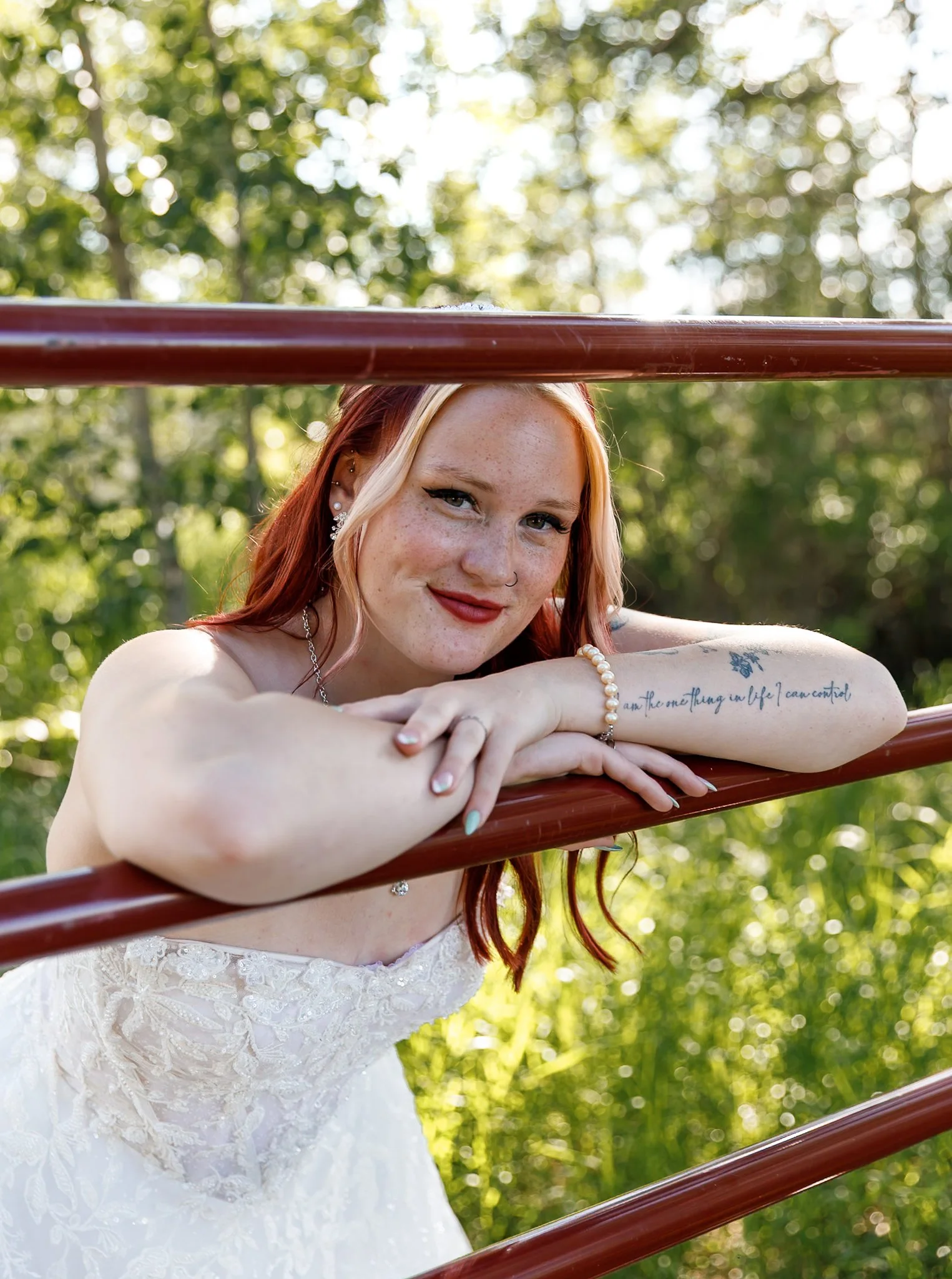 A woman with red hair, freckles, and earrings, leaning on a red railing outdoors, smiling at the camera with a background of green trees and sunlight.