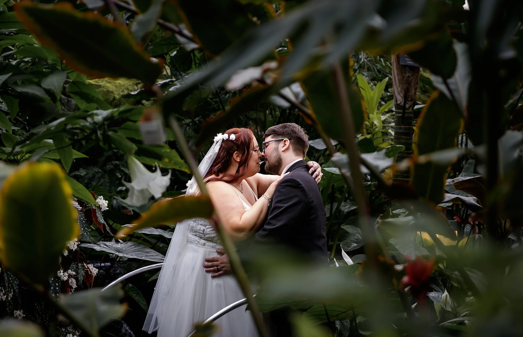 A bride and groom sharing a kiss and intimate moment in a lush green garden with tall tropical plants, one with white flowers, surrounding them.