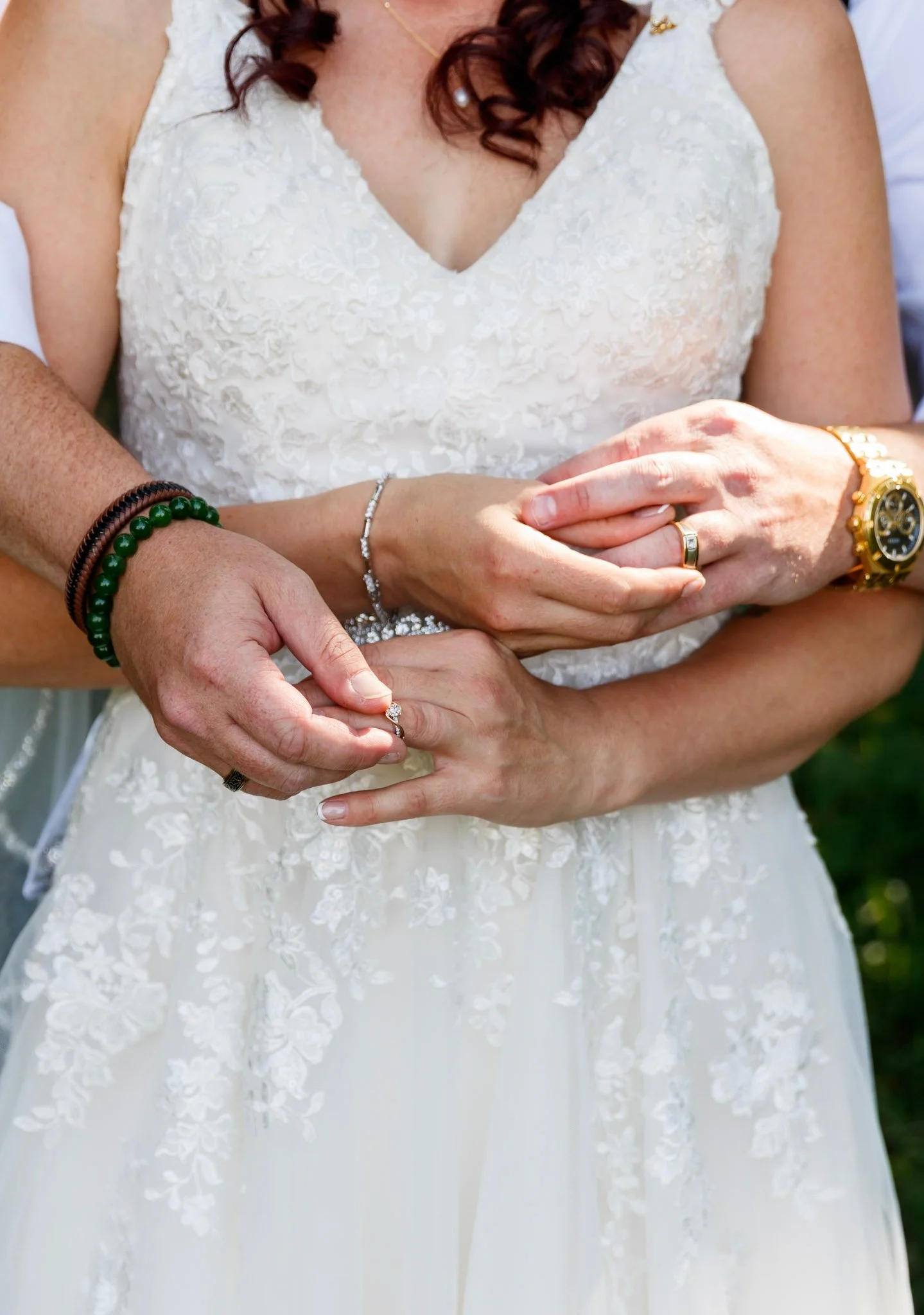 A woman in a white wedding dress is holding her hands together; she is wearing a ring, a wristwatch, a bracelet, and a beaded bracelet. Another person's hands are gently holding hers from behind.