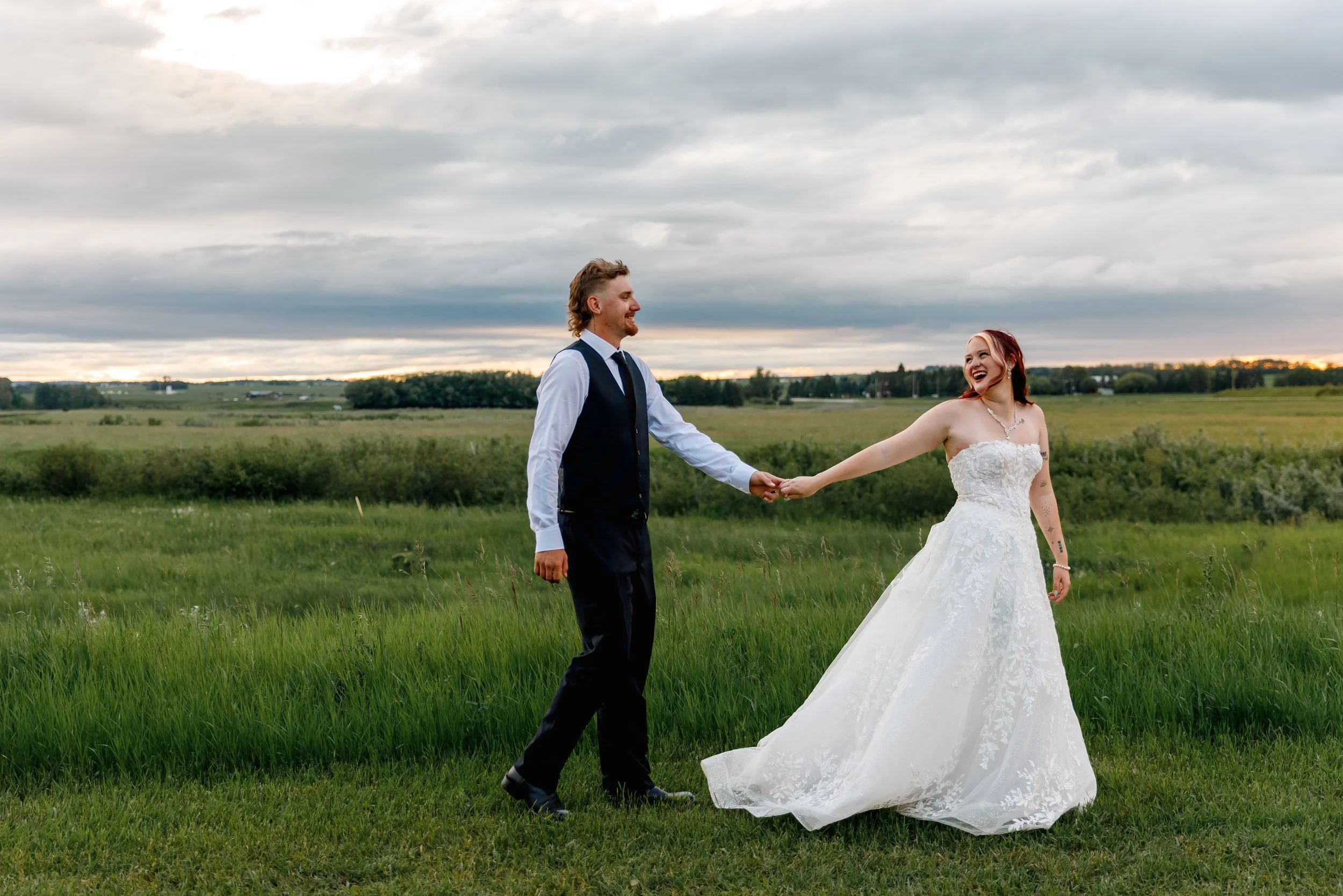 A bride and groom holding hands and smiling in a green field during sunset, with a cloudy sky overhead.