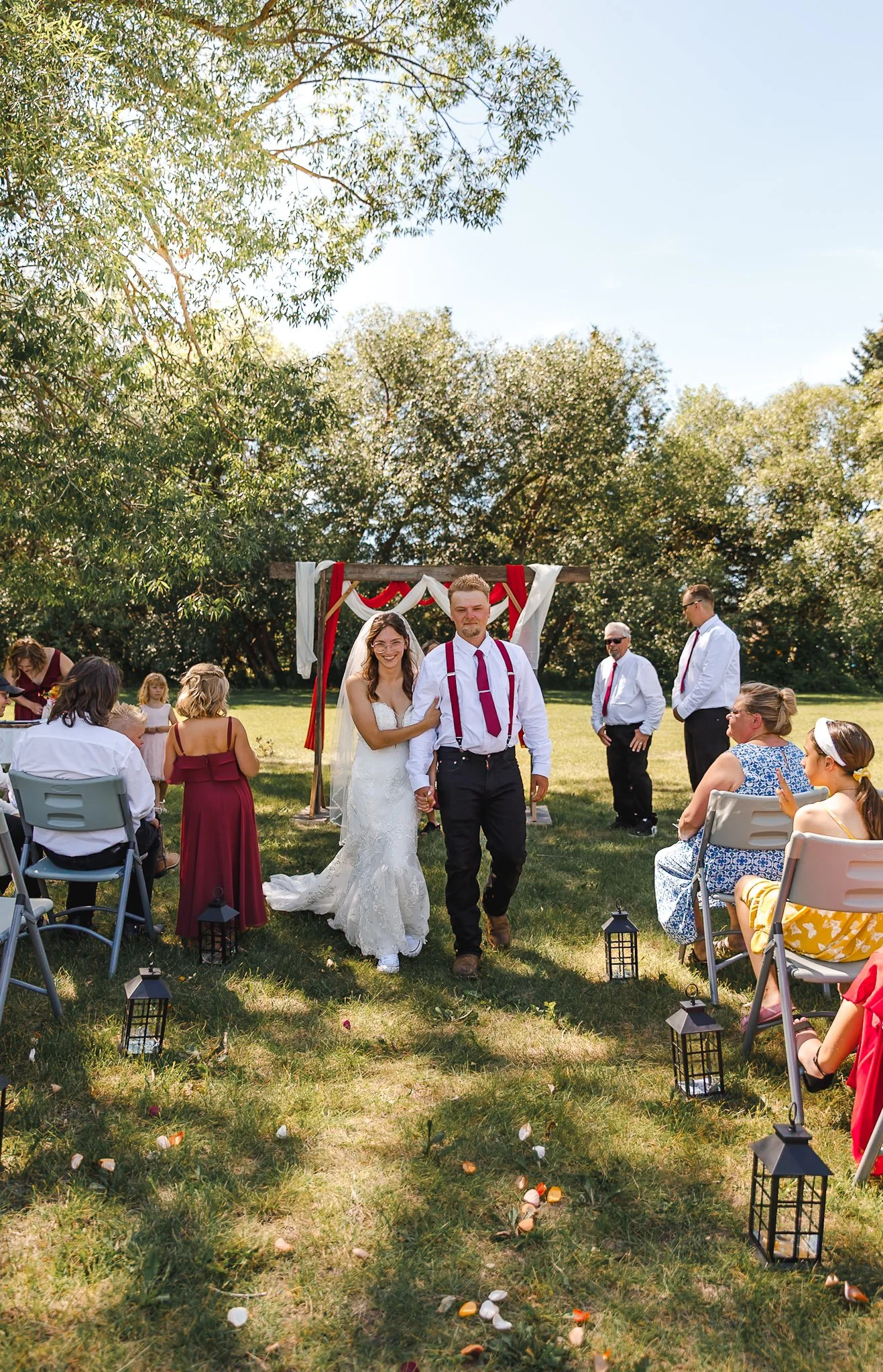 A bride and groom walking down the aisle at an outdoor wedding ceremony on a sunny day, surrounded by seated guests and lantern decorations along the grassy aisle.