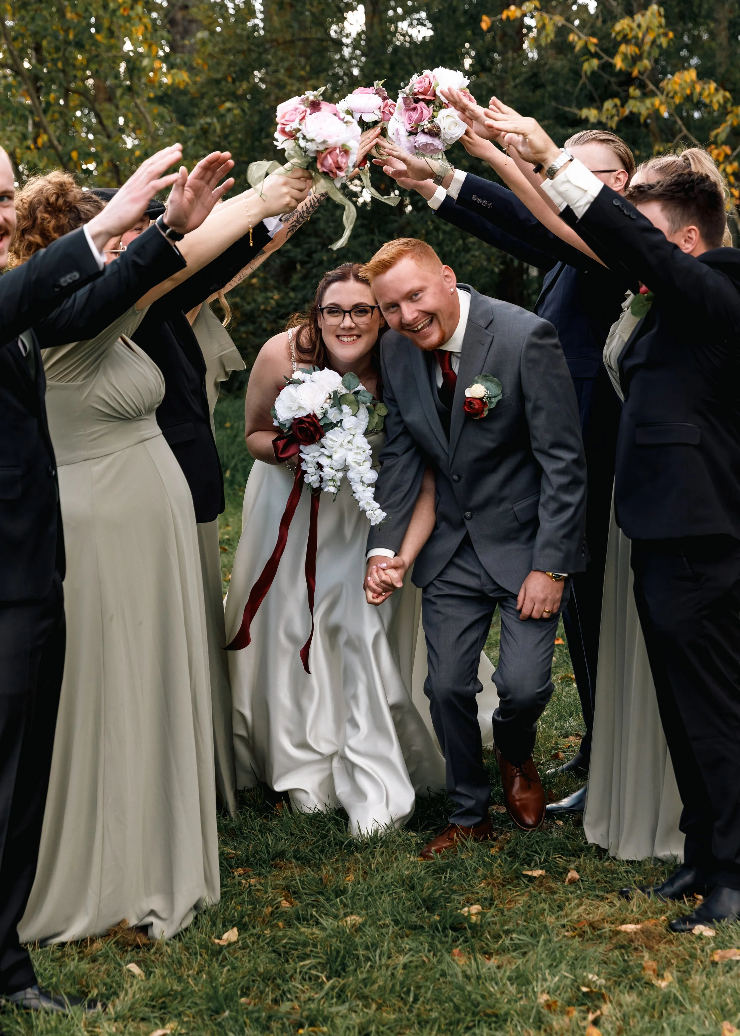 A newlywed couple holding hands under a floral arch created by friends during an outdoor wedding ceremony, with guests reaching out holding bouquets of pink and white flowers.