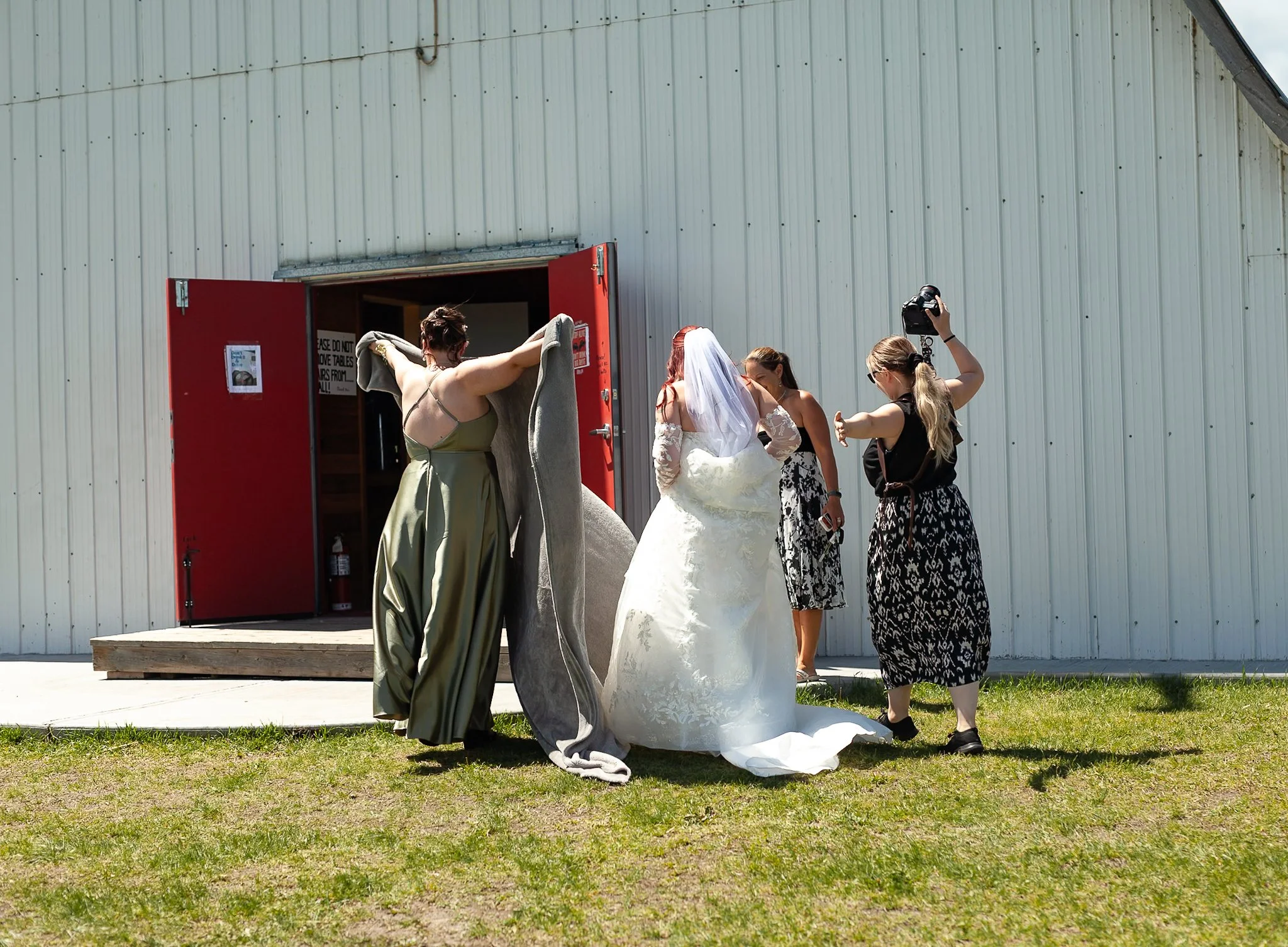 A bride in a white wedding gown and veil With women surrounding her, some adjusting dress or taking photos, outside a barn with a red door and metal siding.