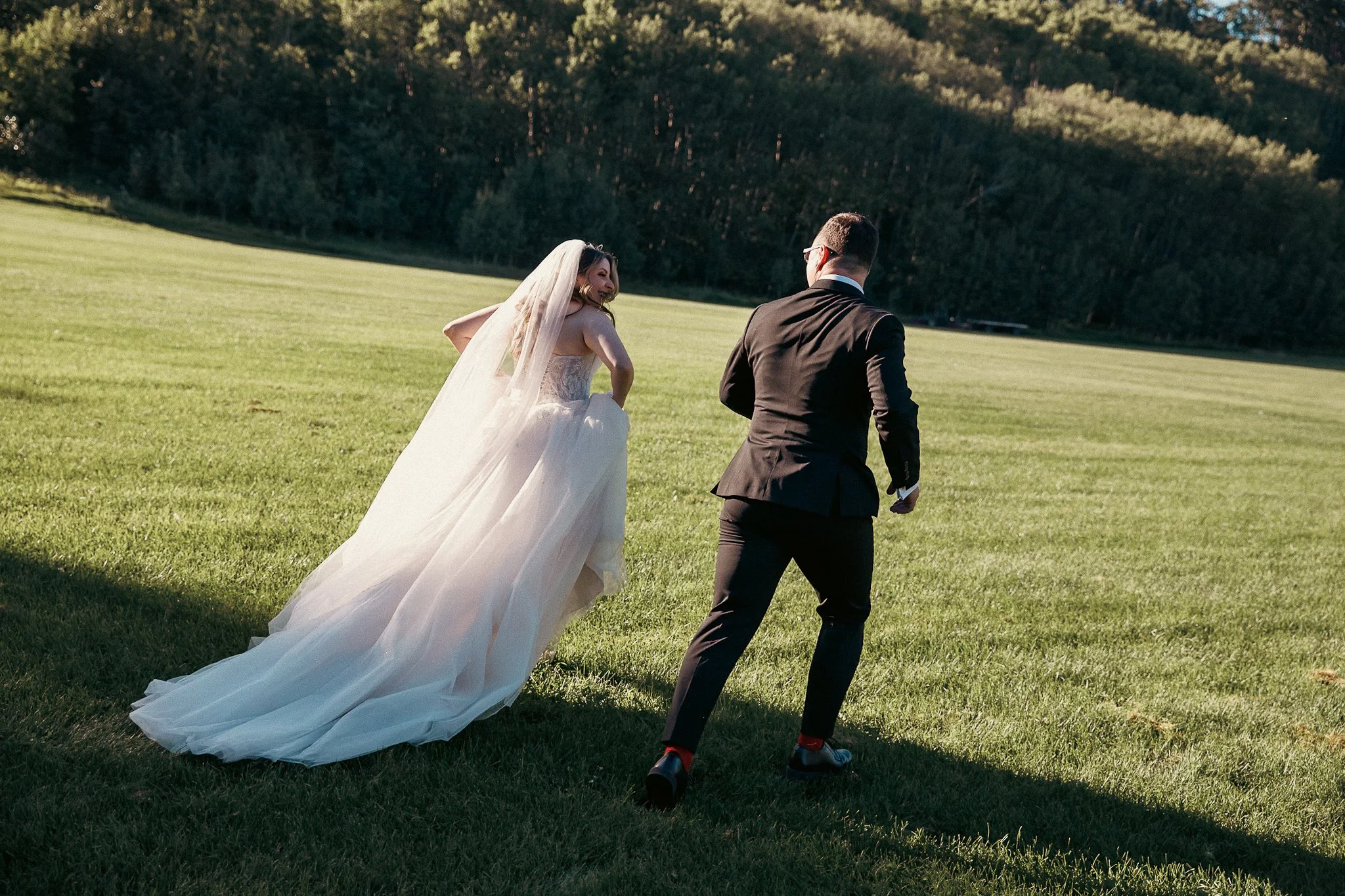 A bride in a white wedding dress and veil smiling behind a groom in a black suit and glasses running outdoors on a grassy field with trees in the background.