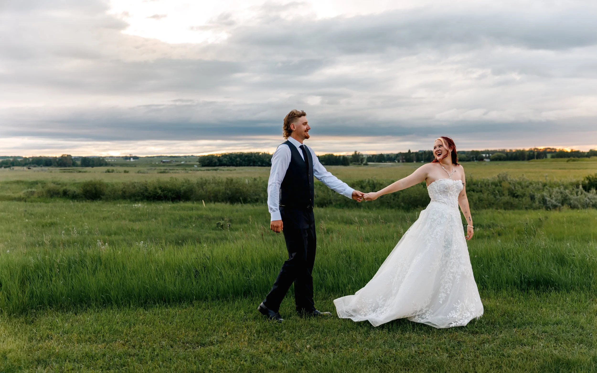 A bride and groom holding hands and smiling in a green field under a cloudy sky during a wedding photoshoot.