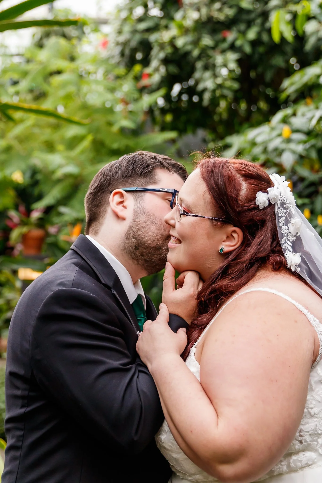 A newlywed couple shares a kiss outdoors, surrounded by green foliage. The man is wearing glasses and a black suit, while the woman has red hair, wears glasses, and a white wedding dress with floral details and a veil.