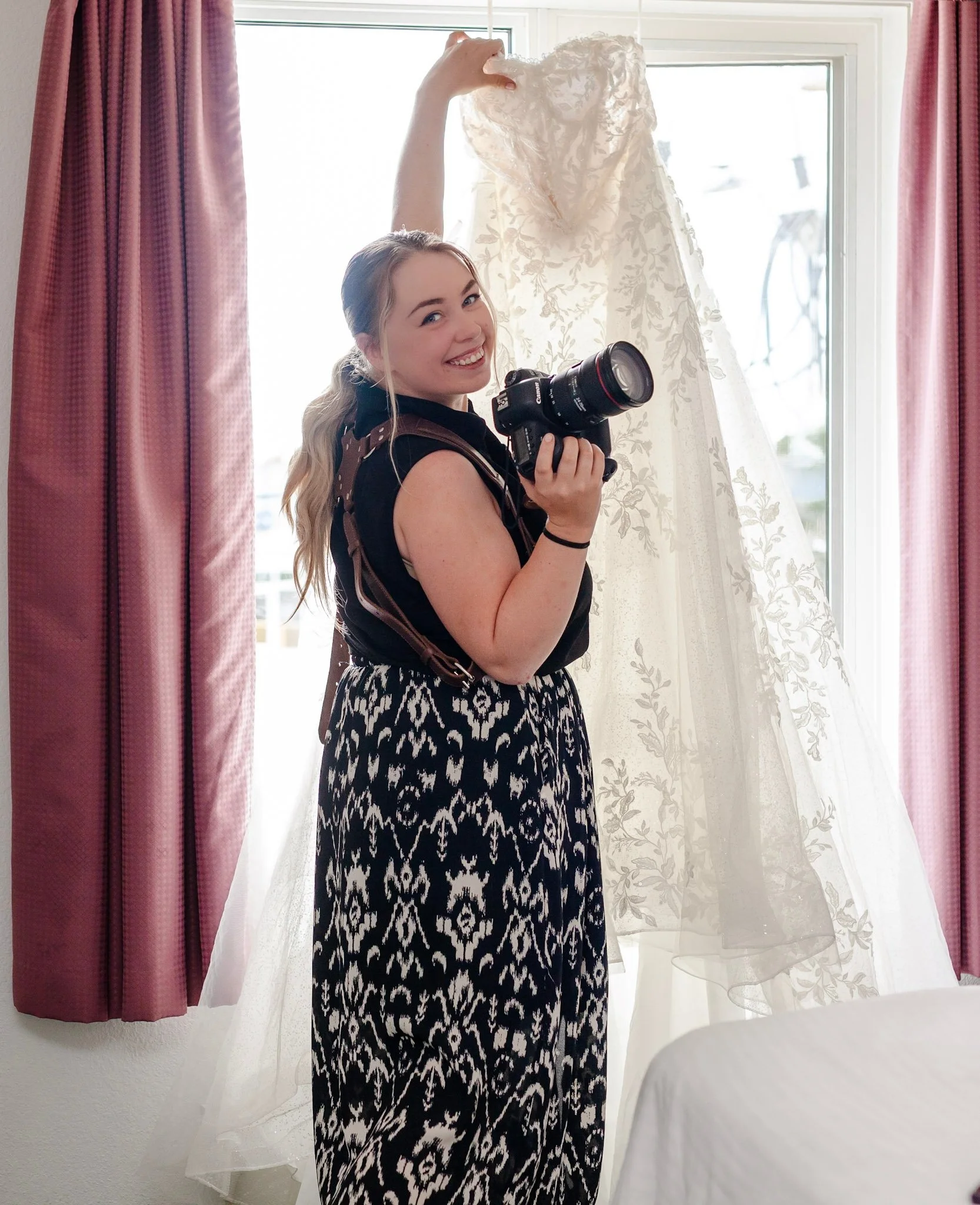 A smiling woman with blond hair holding a camera stands in front of a window with pink and white curtains, wearing a black top and a patterned black and white skirt, holding up a lace dress.