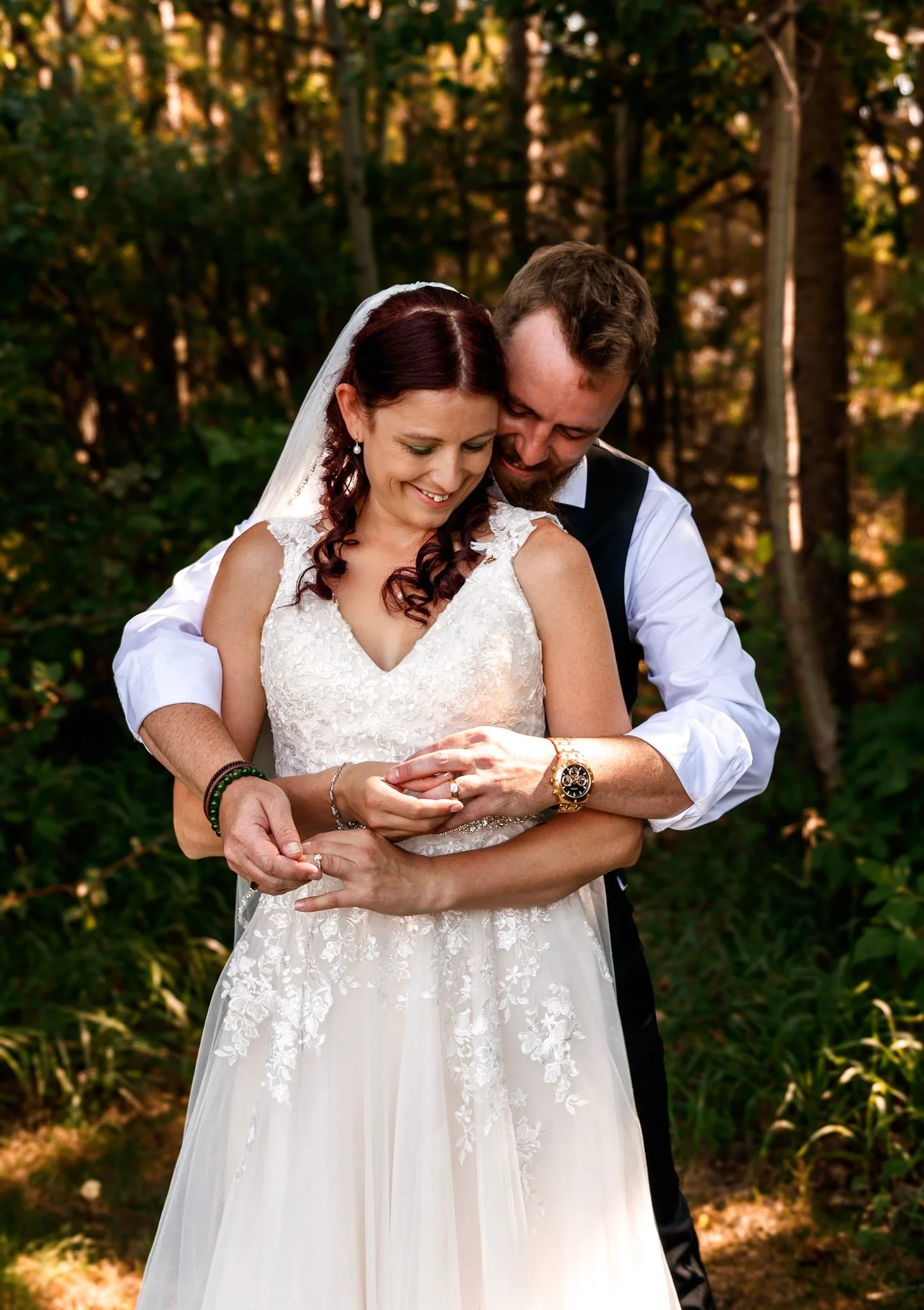 A bride with red hair wearing a white lace wedding dress and a tiara, and a groom with brown hair and a beard wearing a white shirt and black vest, sharing an intimate moment outdoors in a wooded area.