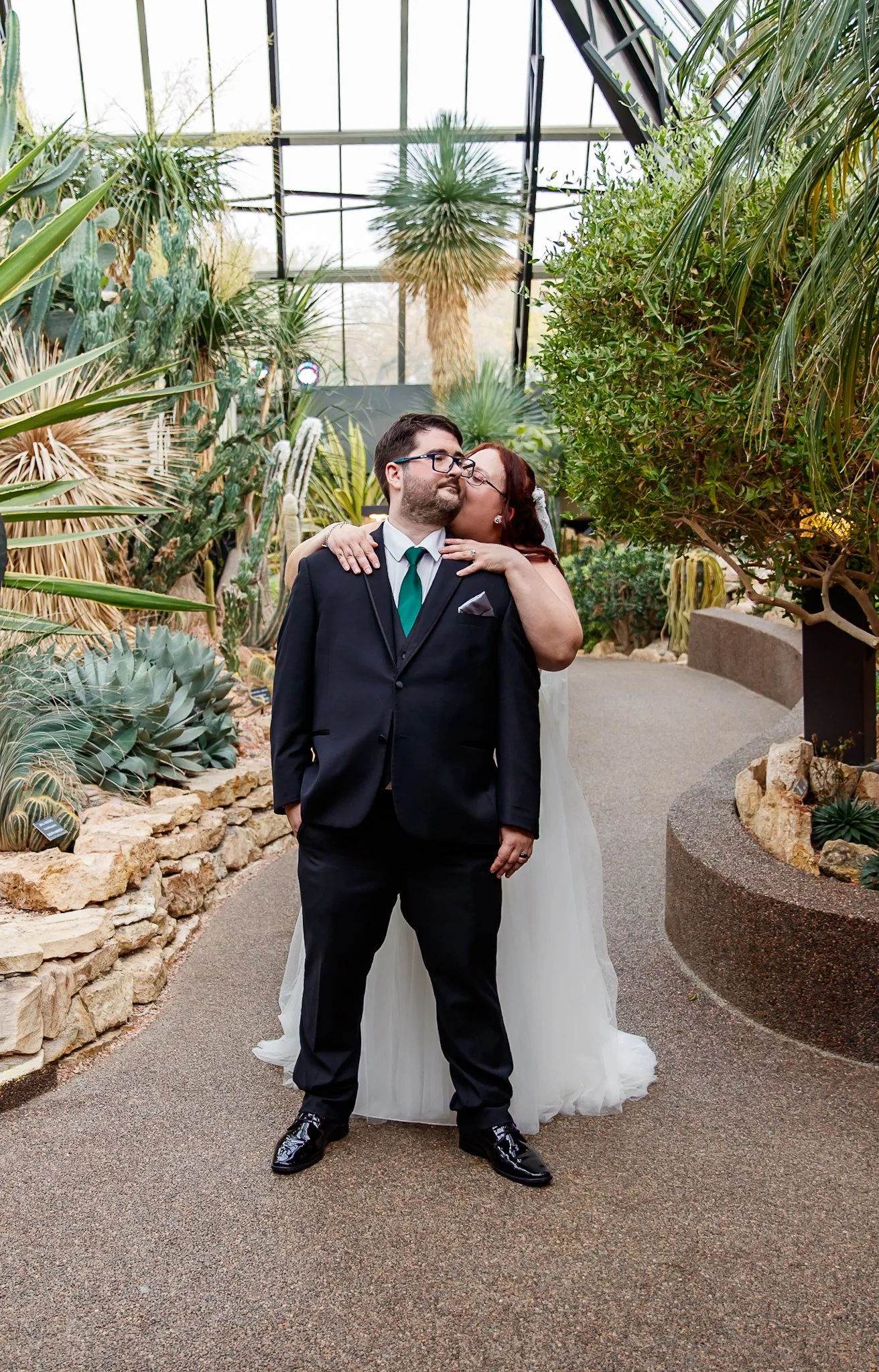A bride kissing a groom on the cheek in a botanical greenhouse. The groom is wearing a dark suit and tie, and the bride is in a white wedding dress. They are surrounded by green plants and succulents.