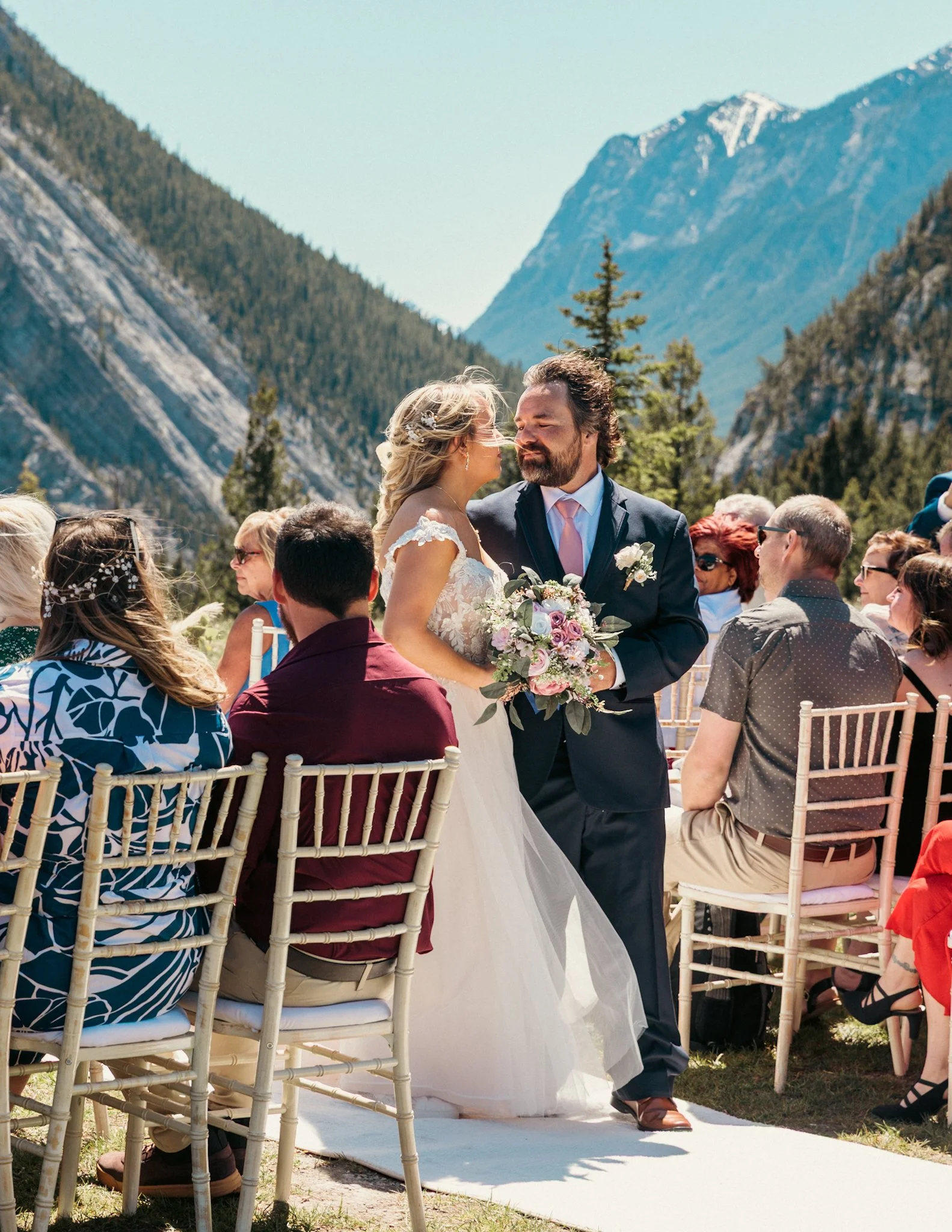 A wedding ceremony taking place at Tunnel Mountain in Banff. The bride and groom are standing under a floral arch, surrounded by guests seated on white chairs on a grassy field. The sky is clear and blue, indicating a sunny day.