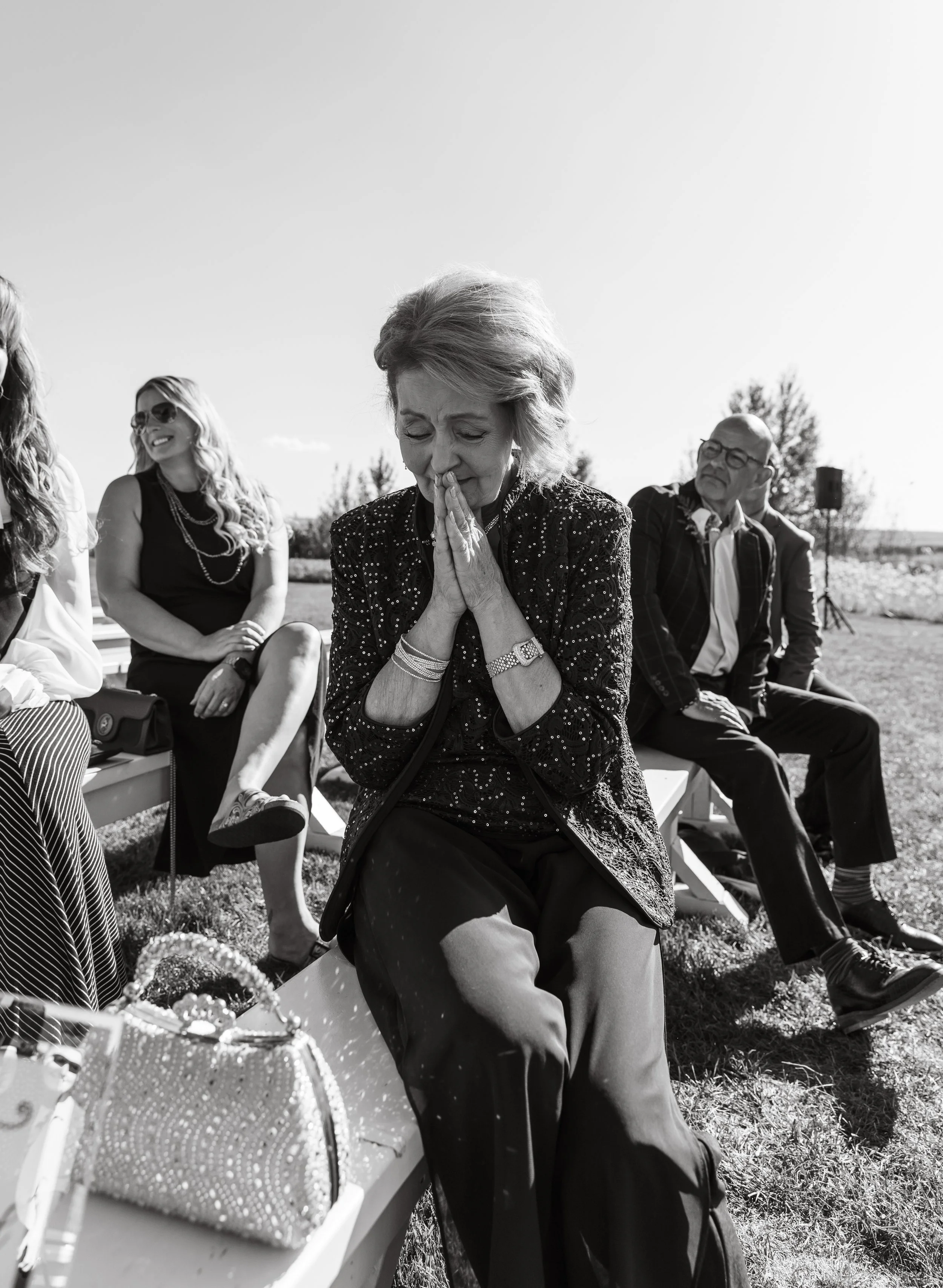 An elderly woman with her hands held in a prayer position, sitting outdoors among other people. She appears emotional or crying. Other adults are smiling and seated nearby, with a clear sky and trees in the background.