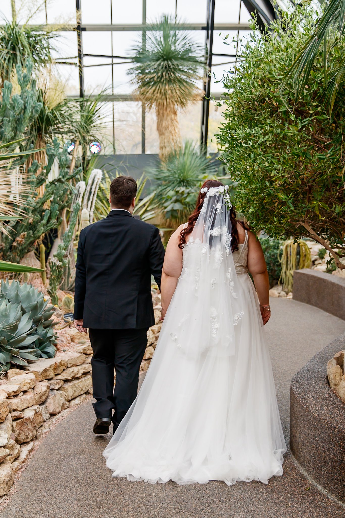 A bride and groom walking together in a greenhouse filled with desert plants. The bride is wearing a white wedding gown with a veil, and the groom is dressed in a black suit. They are holding hands and walking away from the camera.