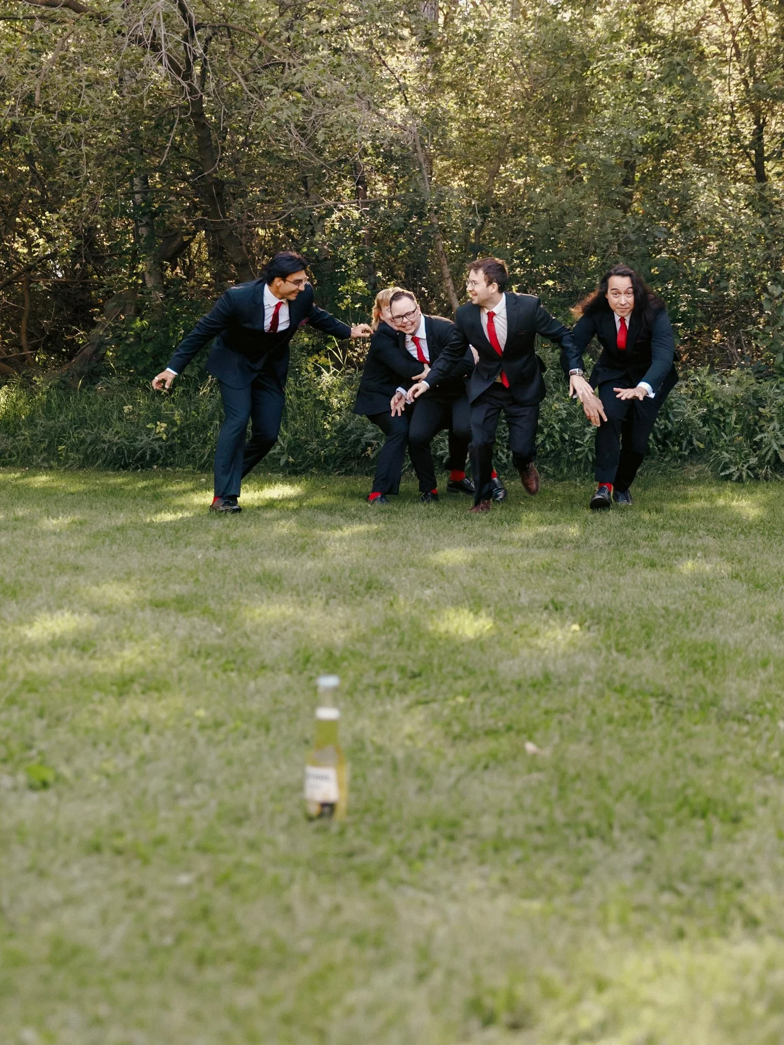 Four people in business suits, red ties, and black shoes are outside near a hedge, bending over and appearing to be in a playful or joking pose around a small stack of white cups on the grass.