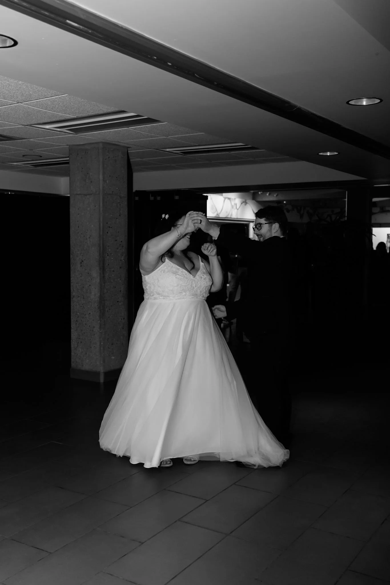 A bride and groom dancing together at a wedding reception. The bride is wearing a white wedding dress and the groom a dark suit, both smiling and holding hands.