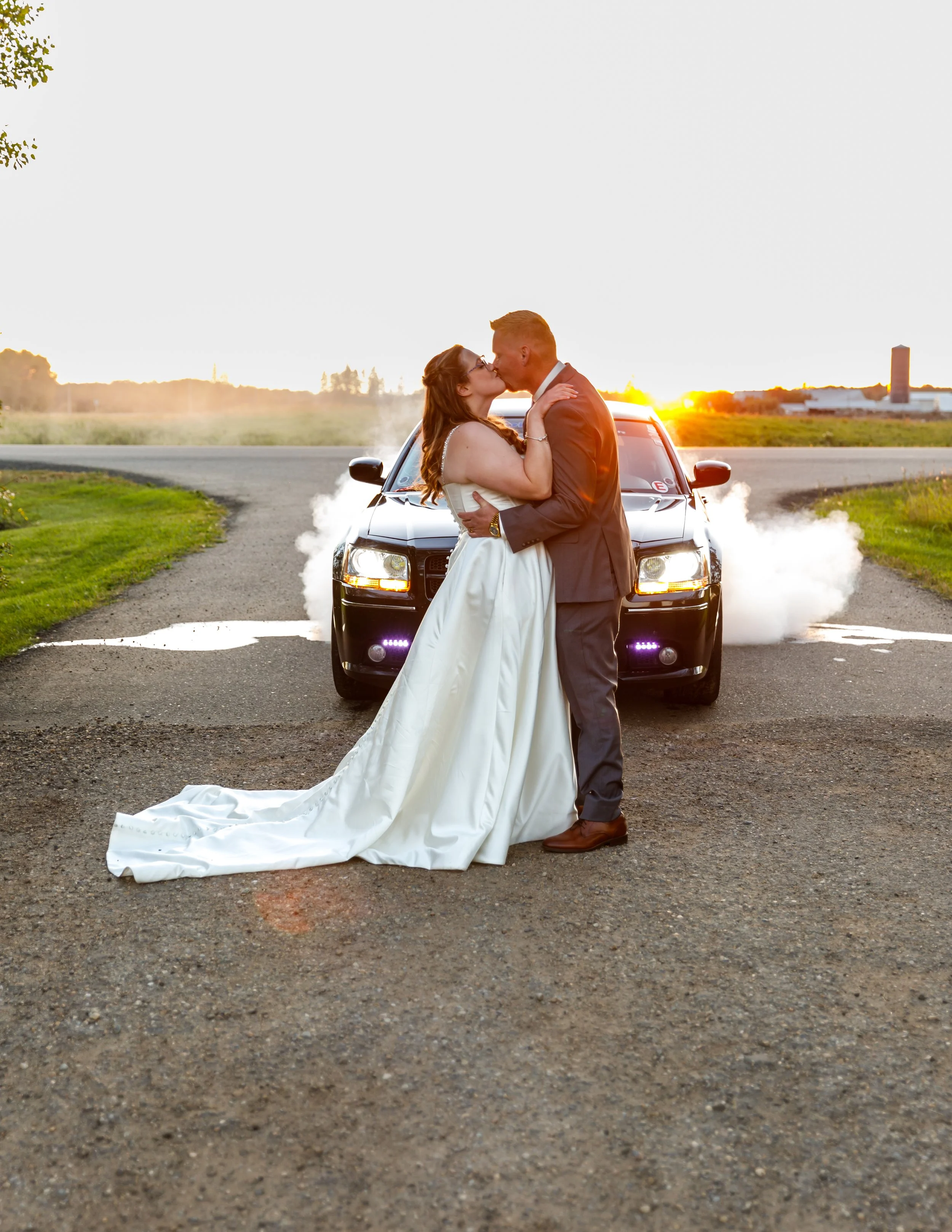 A bride and groom sharing a kiss in front of a black car with exhaust smoke and a sunset in the background.