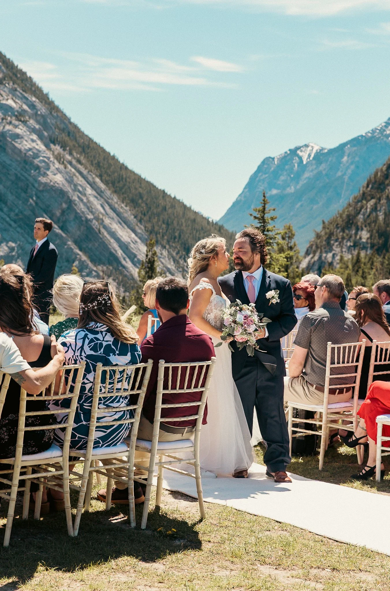 A bride and groom sharing a kiss at their outdoor wedding ceremony surrounded by guests, with mountains and blue sky in the background.