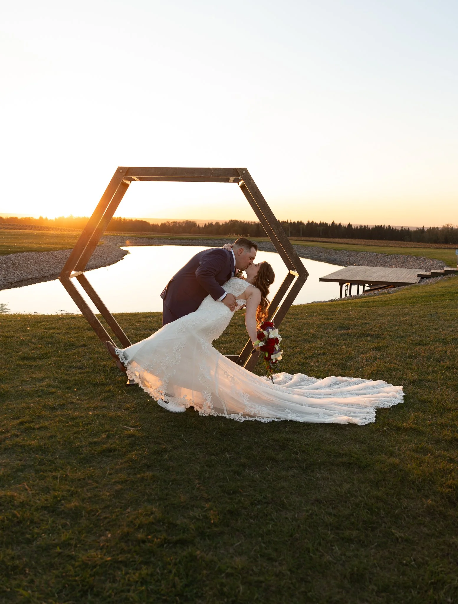 A couple in wedding attire sharing a kiss under a geometric wooden arch at sunset near a lake with lush greenery at Tin Roof Event Center in Lacombe, Alberta