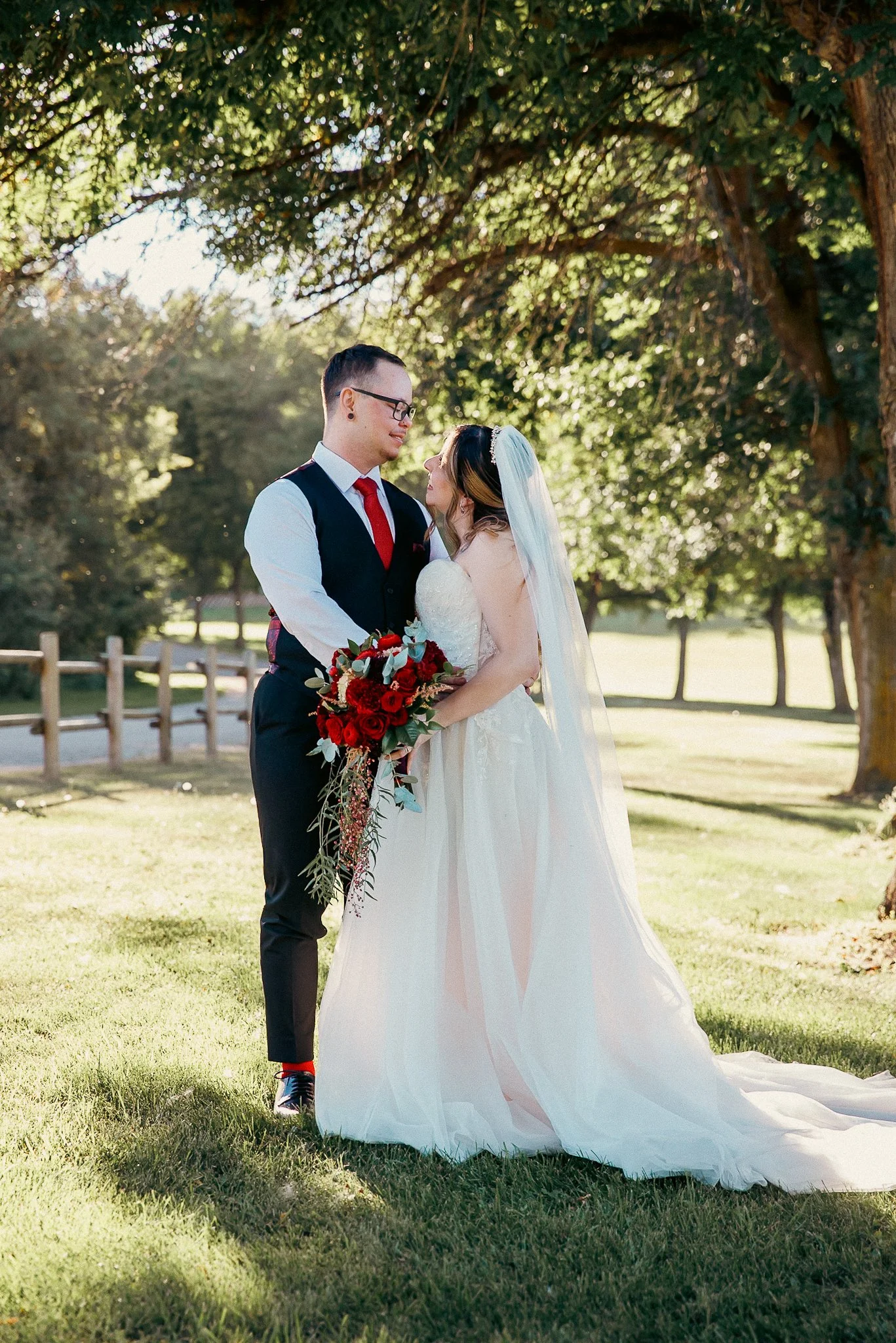 A bride and groom standing outdoors under a large leafy tree during daytime, holding a bouquet of red flowers, with a wooden fence and a field in the background.