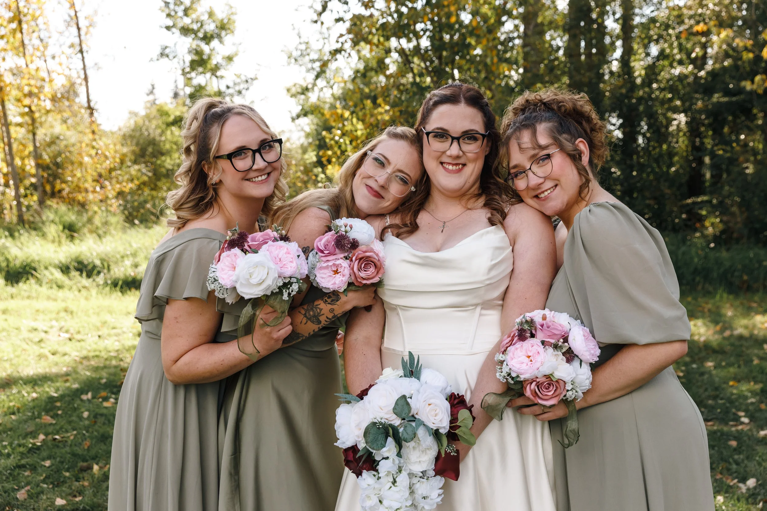 A group of four women smiling together outdoors during a celebration, with trees and sunlight in the background. The women are holding bouquets of flowers and wearing dresses in neutral tones.