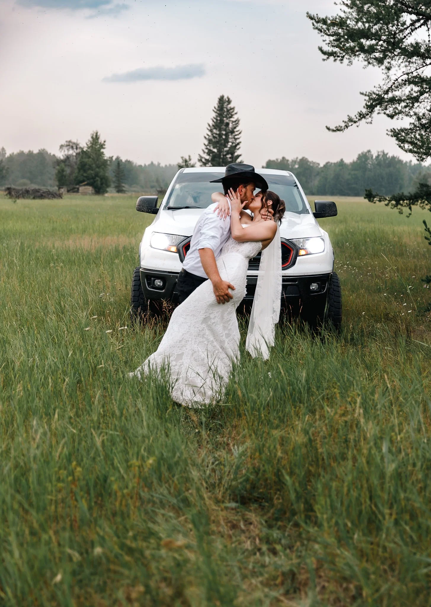 A couple kissing in a field with a white pickup truck behind them, surrounded by green grass and trees.