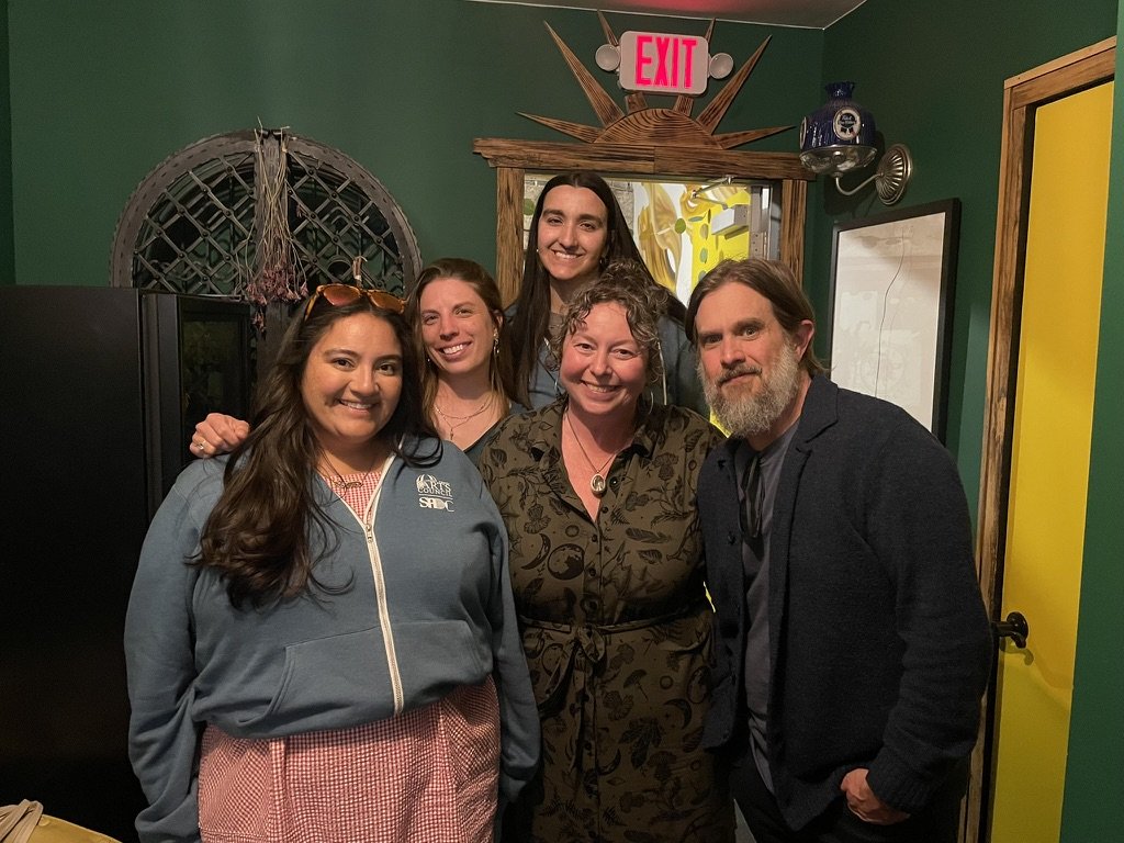 Group of five people smiling and posing together indoors with green walls, a yellow door, and decorative items in the background.