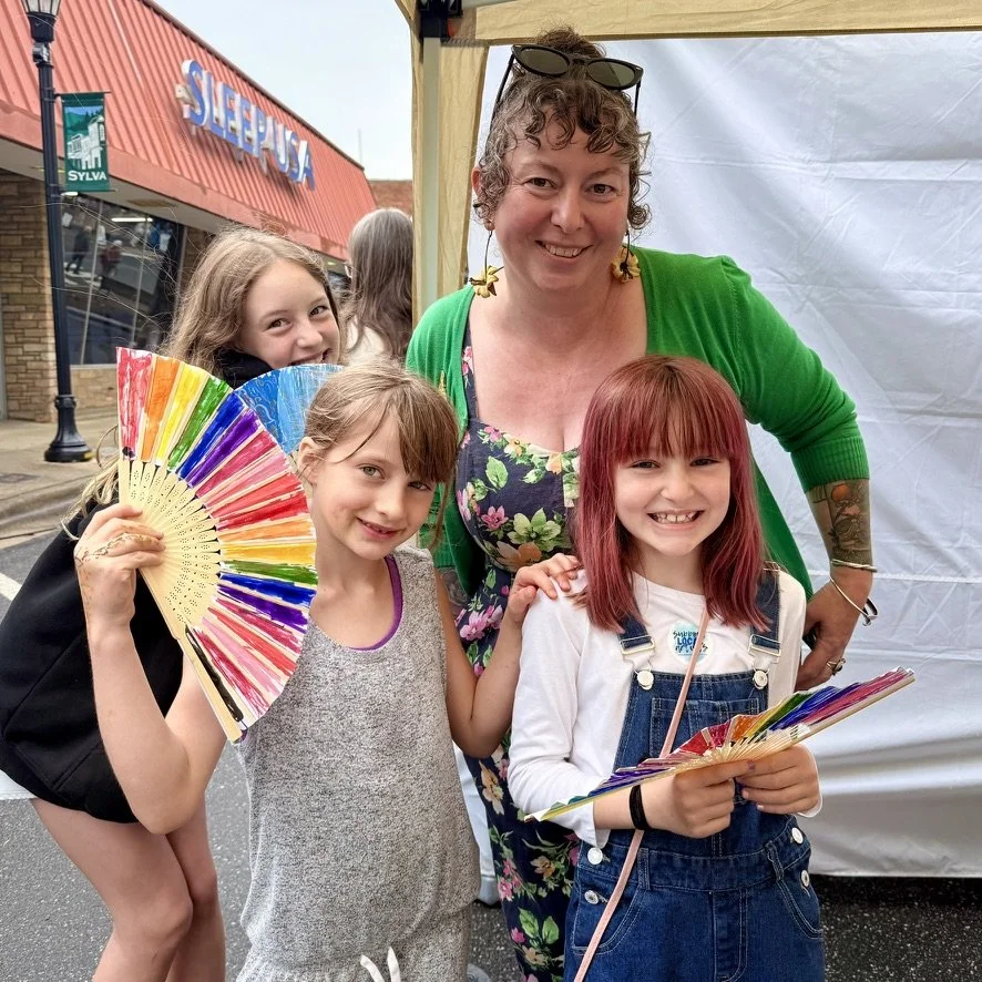 A woman with curly hair and sunglasses on her head, wearing a green cardigan and floral dress, poses with three young girls. One girl with blonde hair and a black backpack holds a colorful rainbow fan, another girl with brown hair in a gray dress also holds a rainbow fan, and the third girl with red hair and overalls holds a rainbow fan. They are outdoors near a storefront with a sign that says "SLEEPY'S."