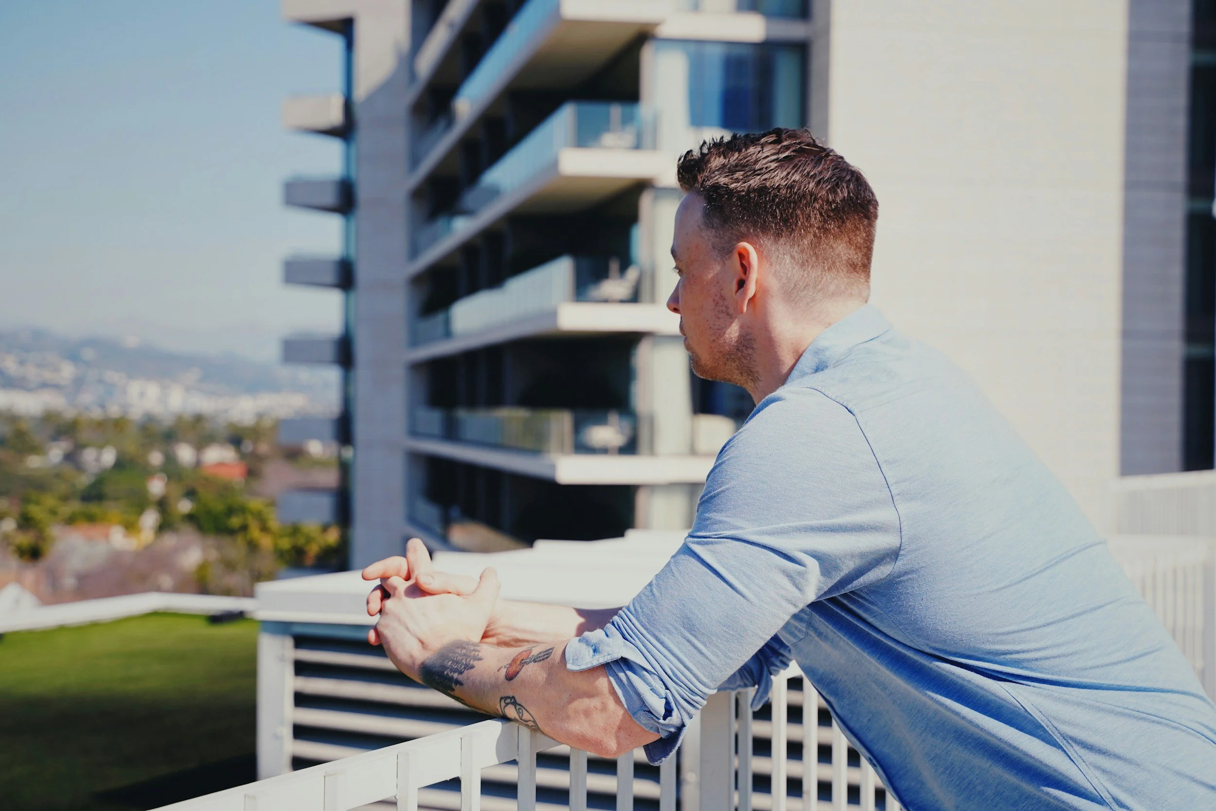 A man with tattoos on his right arm, wearing a light blue shirt, is leaning on a white balcony railing, looking to the right. In the background, there is a modern building with multiple balconies and large windows, and a distant cityscape with hills.