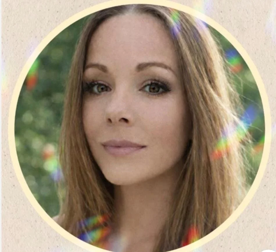 A woman with light brown hair and makeup, outdoors with blurred greenery in the background.