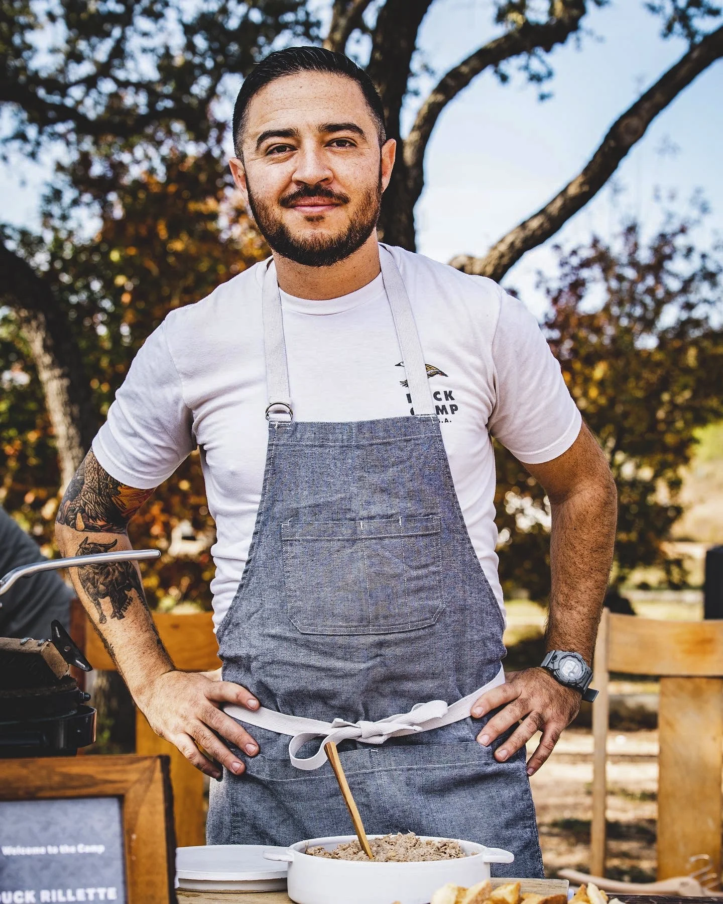A man with a beard and tattoos wearing a white t-shirt, gray apron, and a watch, standing outdoors with hands on hips, smiling, with trees and a wooden fence in the background, cooking or serving food.