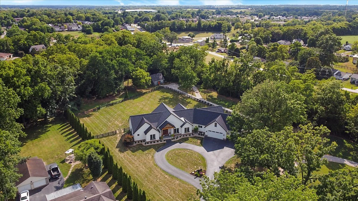 Aerial view of a large house with a circular driveway, surrounded by trees and a fenced yard, in a suburban neighborhood.