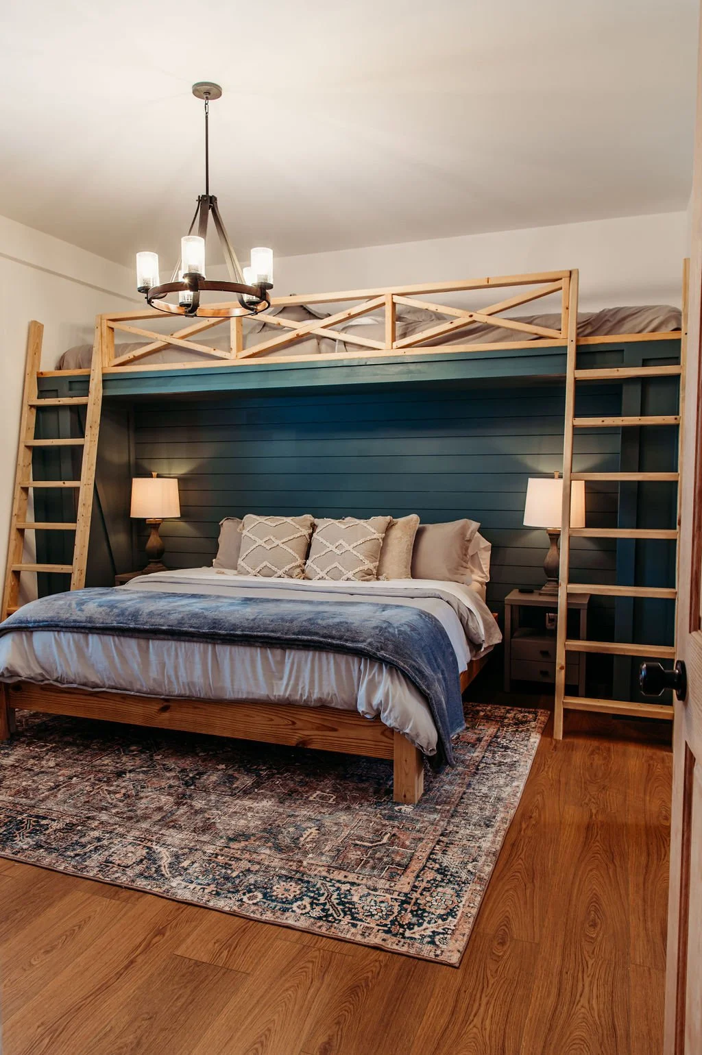 A bedroom with a double bed against a blue paneled wall, a loft bed above with wooden railings and ladders on each side, and a chandelier hanging from the ceiling. The double bed has beige and patterned pillows, a blanket, and is flanked by two night