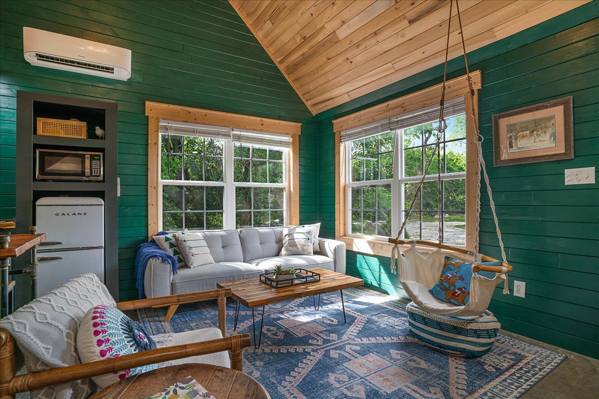 Cozy living room with green shiplap walls, wooden ceiling, large windows, white sofa, colorful throw pillows, wooden coffee table, hanging swing chair, blue patterned rug, mini fridge, microwave, air conditioner, and framed painting.