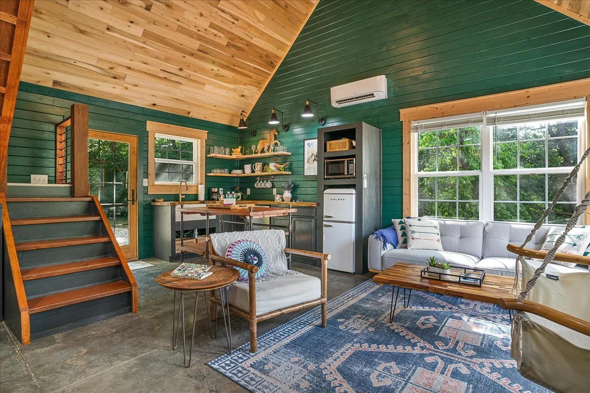 Interior of a cozy living space with green and wood-paneled walls, a sloped ceiling, a white sofa, a small wooden table, a hanging swing chair, and a kitchen area with open shelves, a mini refrigerator, microwave, and an air conditioning unit.