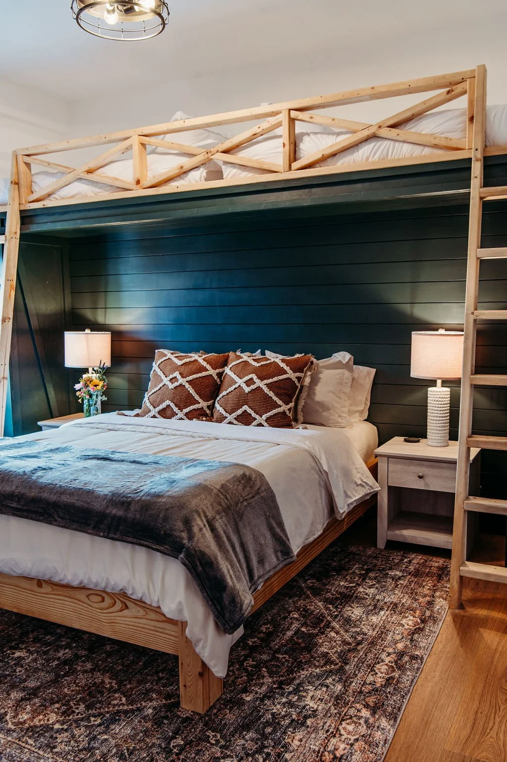 Bedroom with a wooden loft bed featuring a top bunk with bedding and a safety railing, two bedside tables with lamps, a rug, and decorative pillows.