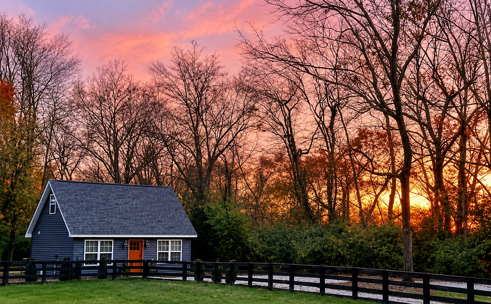 A small blue house with a brown door and white-framed windows is situated in front of a lush green lawn, with a black fence around it. In the background, a dense line of leafless trees is visible against a colorful sunset sky with shades of pink, ora