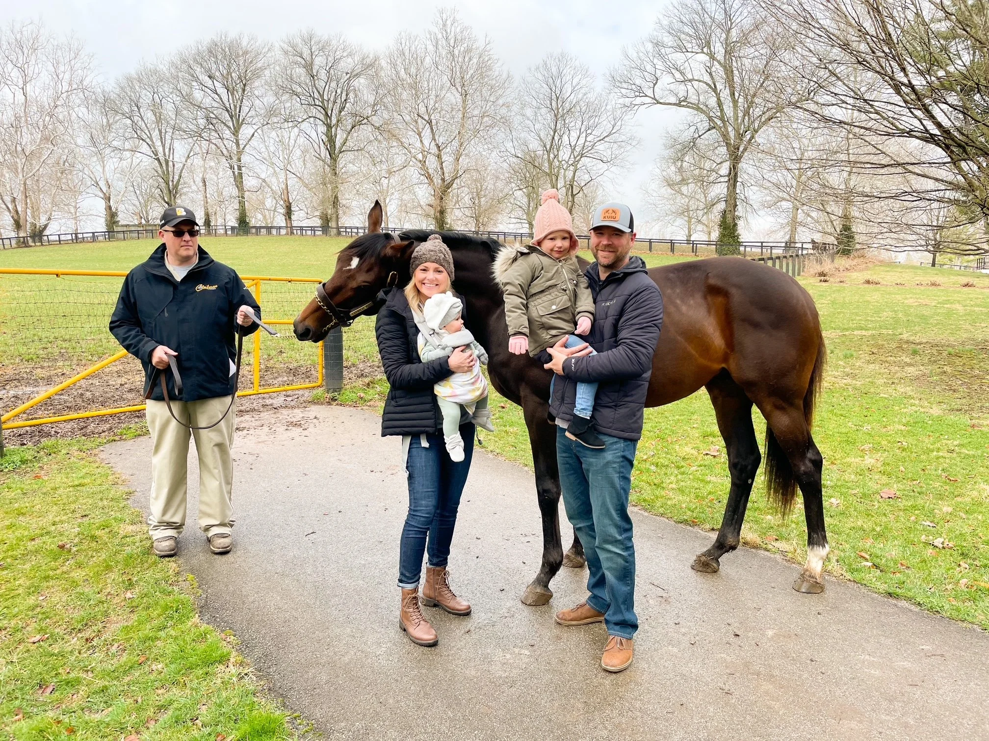 Family of four with two young girls and two men standing near a horse on a wet paved path in a park with leafless trees in the background, overcast sky.